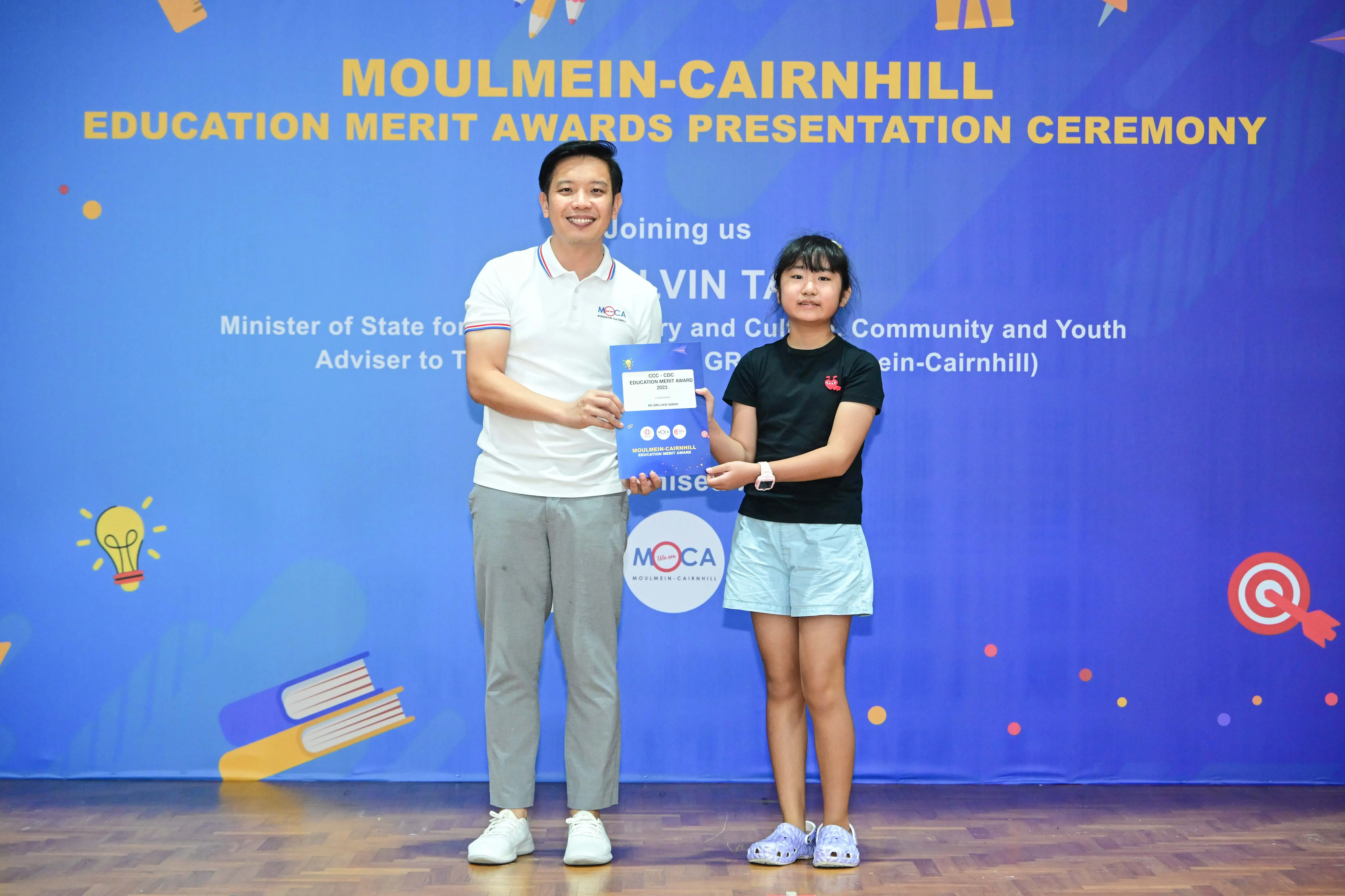 A man and a young girl hold a certificate together at the Moulmein-Cairnhill Education Merit Awards ceremony.