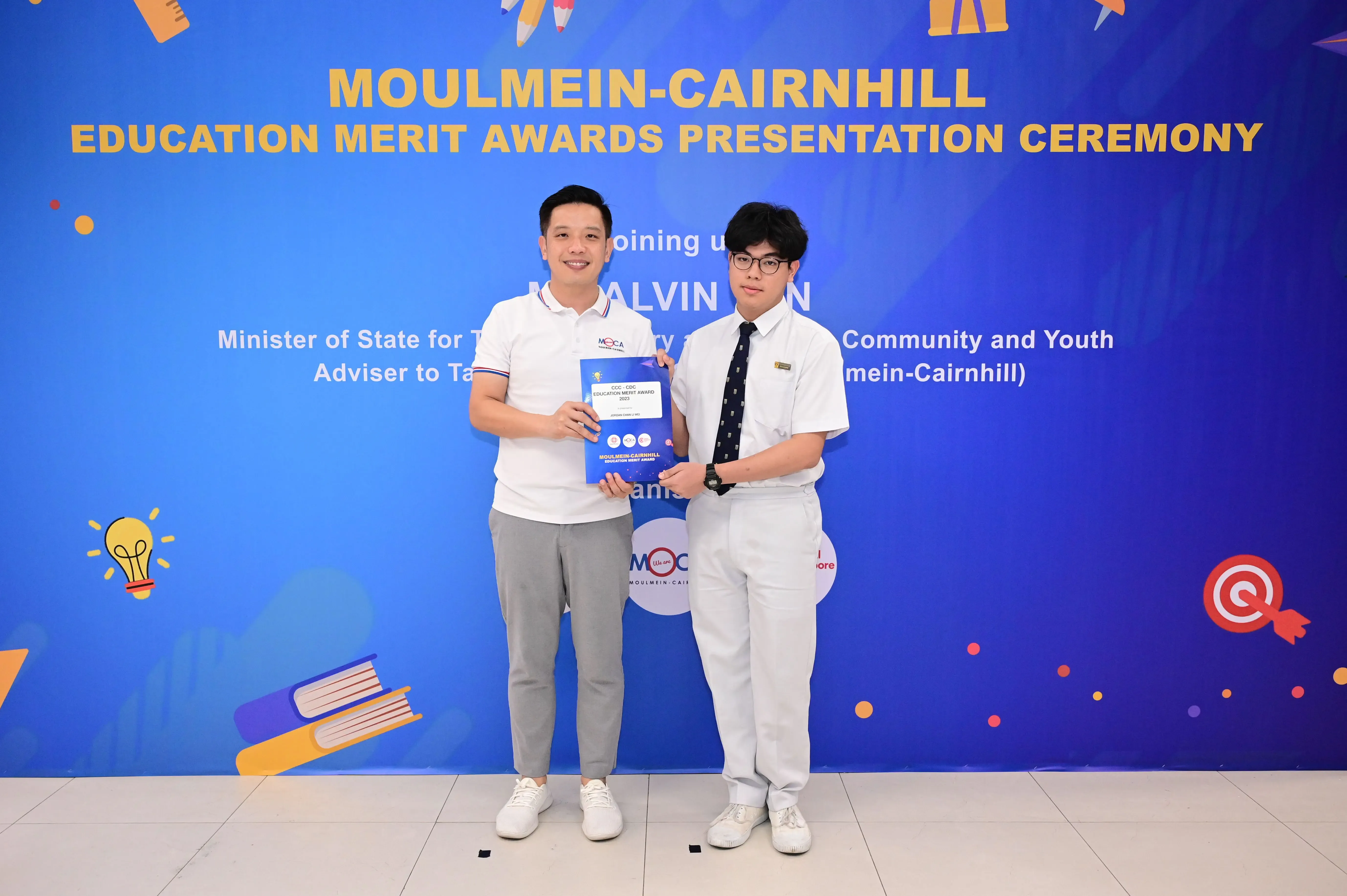 Two men hold a certificate together in front of a Moulmein-Cairnhill Education Merit Awards Presentation Ceremony banner.