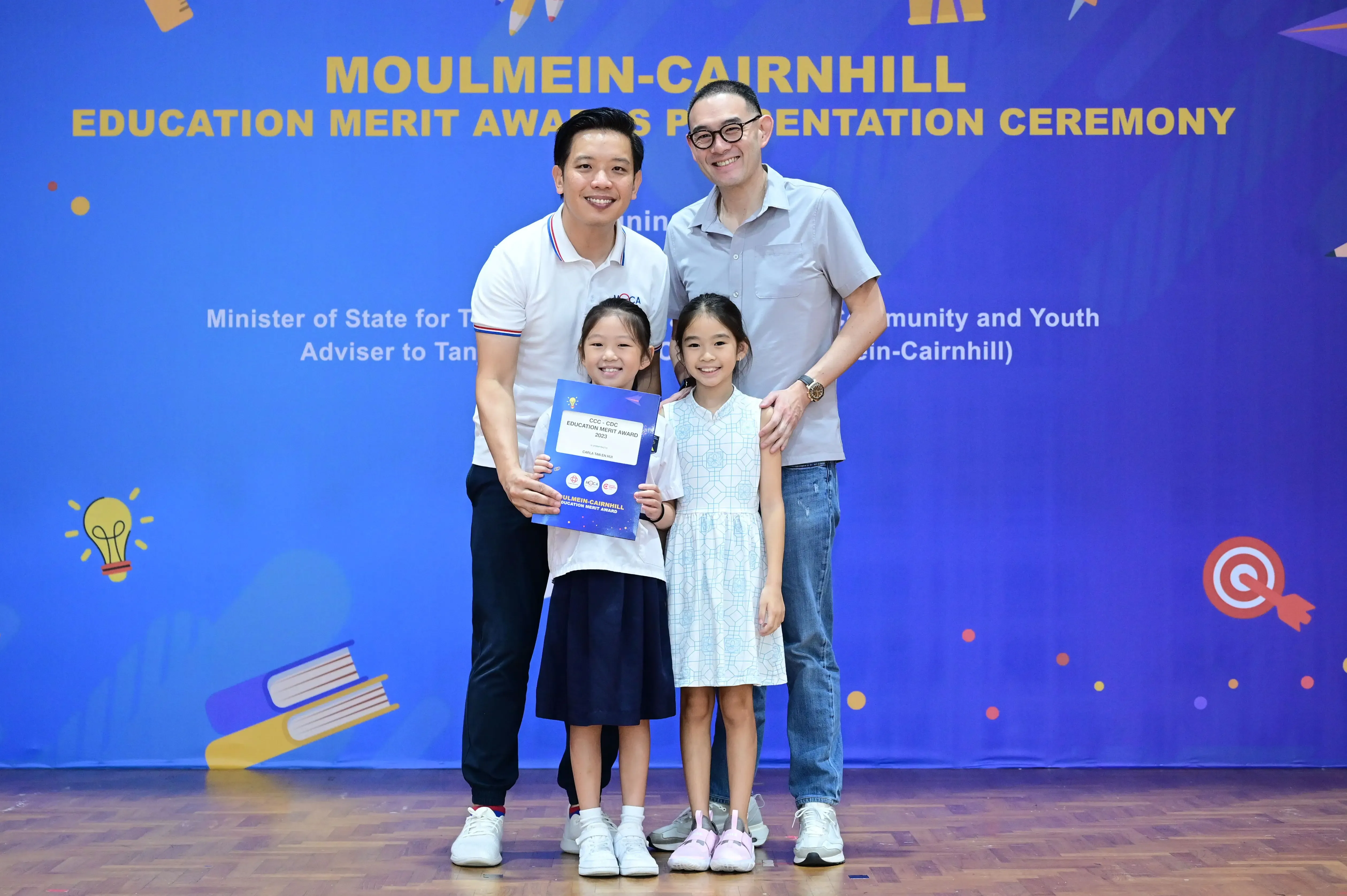 Two men and two girls pose at the Moulmein-Cairnhill Education Merit Awards Presentation Ceremony, one girl holding an award folder.
