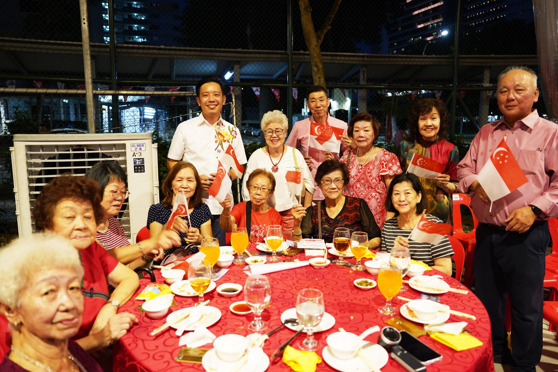 Group of people smiling at a red table outdoors at night, holding small Singapore flags with drinks and dishes nearby.