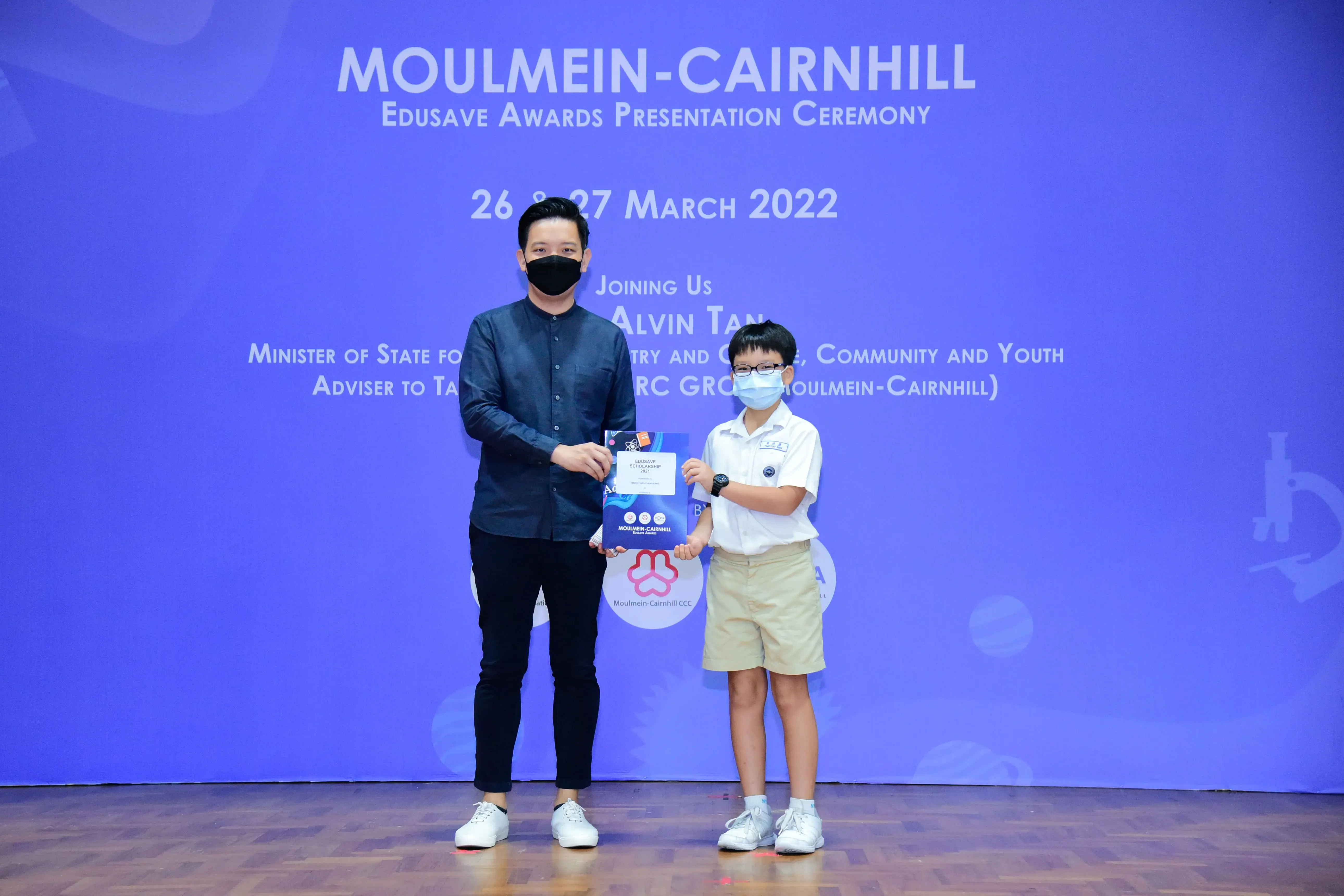 An adult and a young student in school uniform hold an award at the Moulmein-Cairnhill Edusave ceremony, March 2022.
