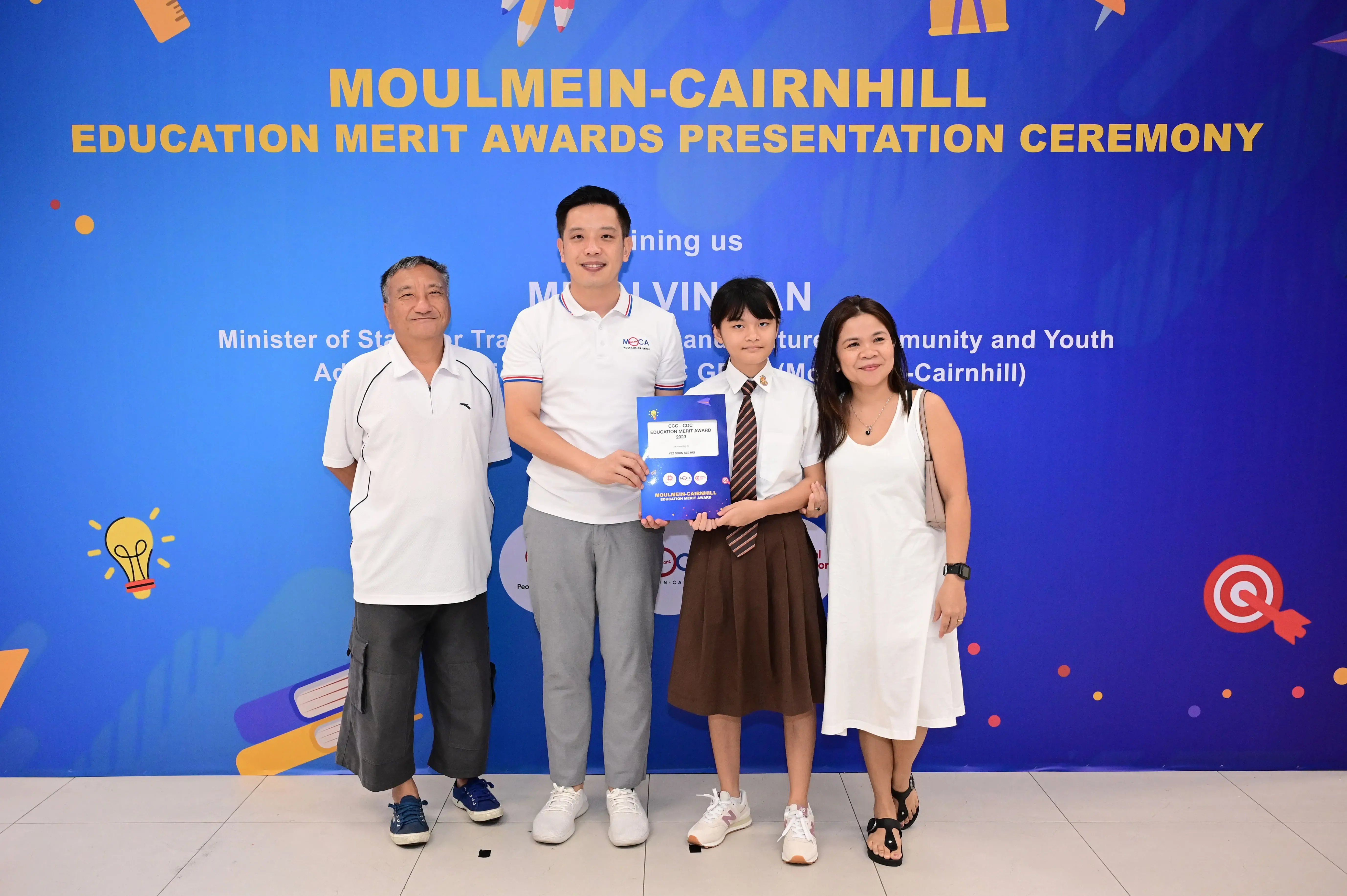 Four people pose holding a certificate in front of a Moulmein-Cairnhill Education Merit Awards banner.