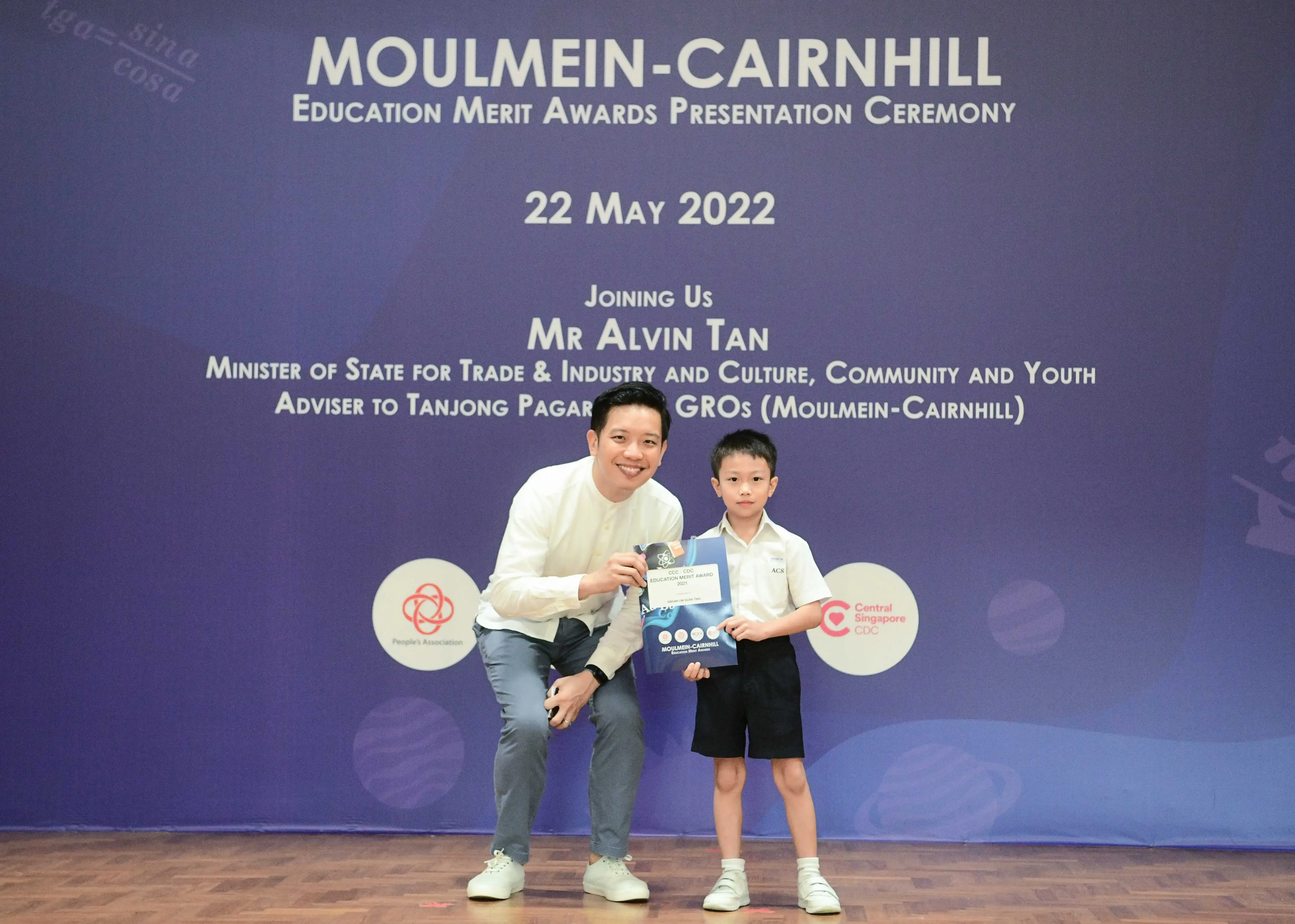 A man crouching beside a young boy holding an award certificate at the Moulmein-Cairnhill Education Merit Awards on 22 May 2022.