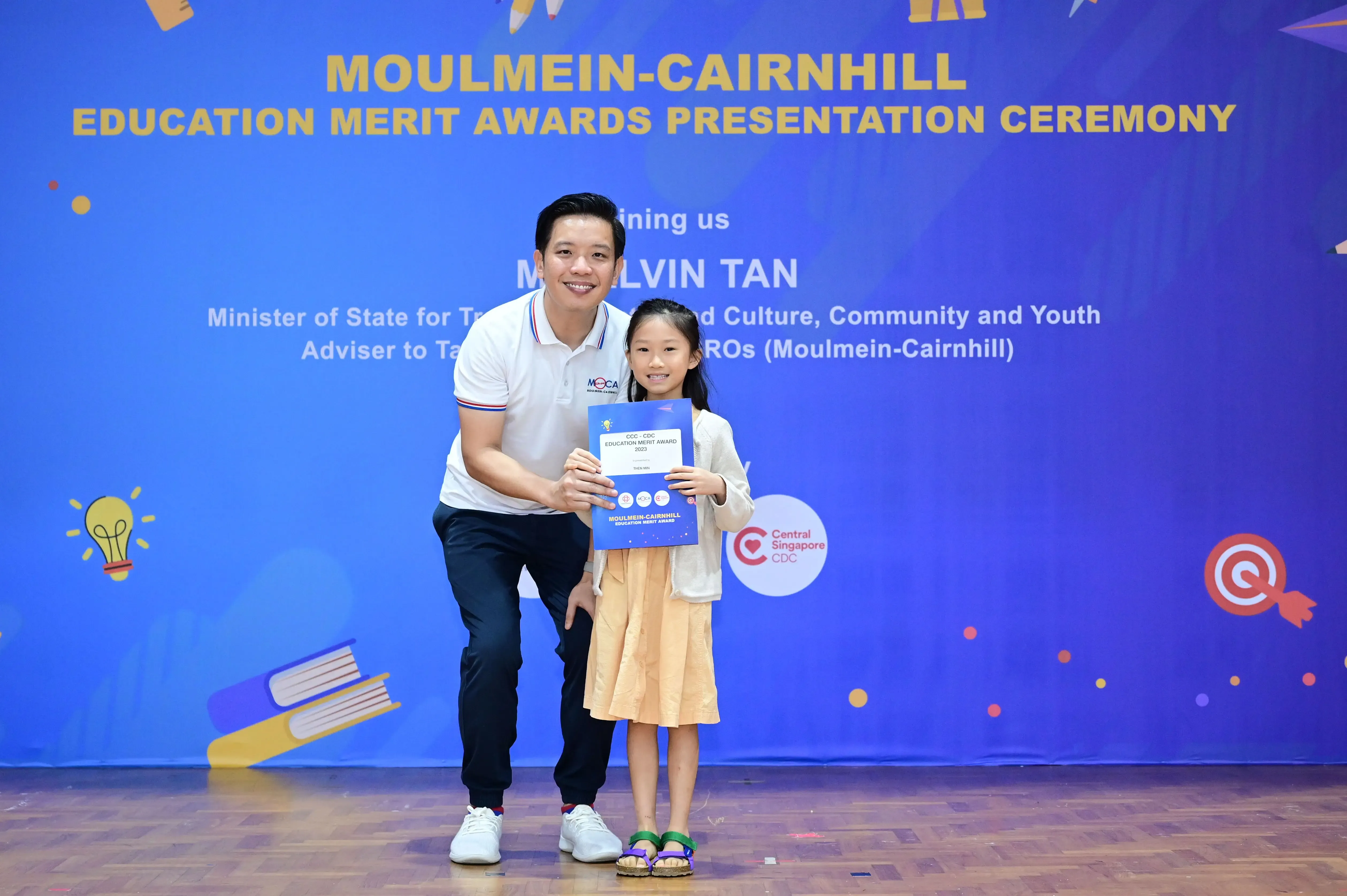 A man and a young girl hold a blue award certificate at the Moulmein-Cairnhill Education Merit Awards Ceremony.