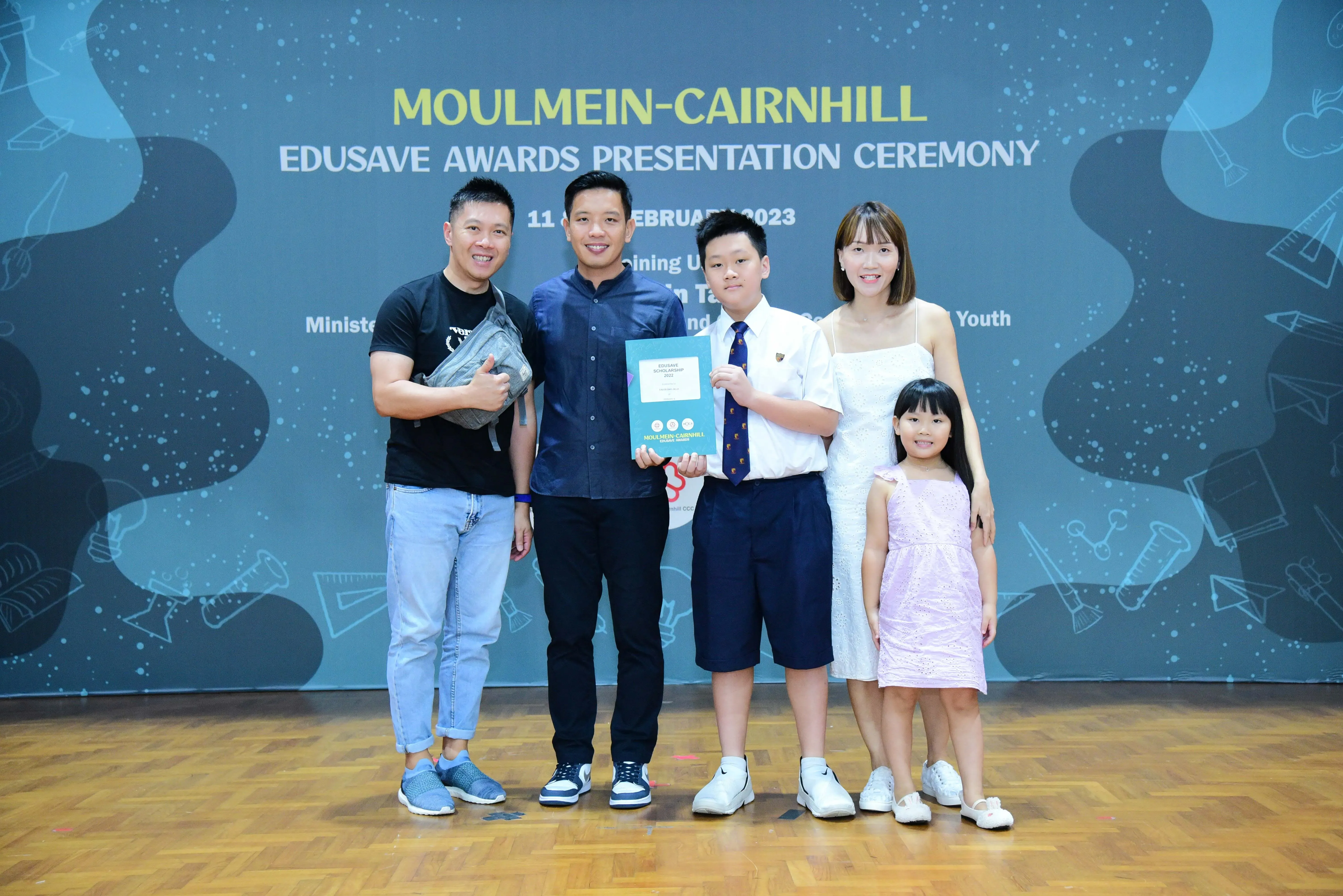 Five people pose at the Moulmein-Cairnhill Edusave Awards Presentation Ceremony, with a boy holding a certificate.