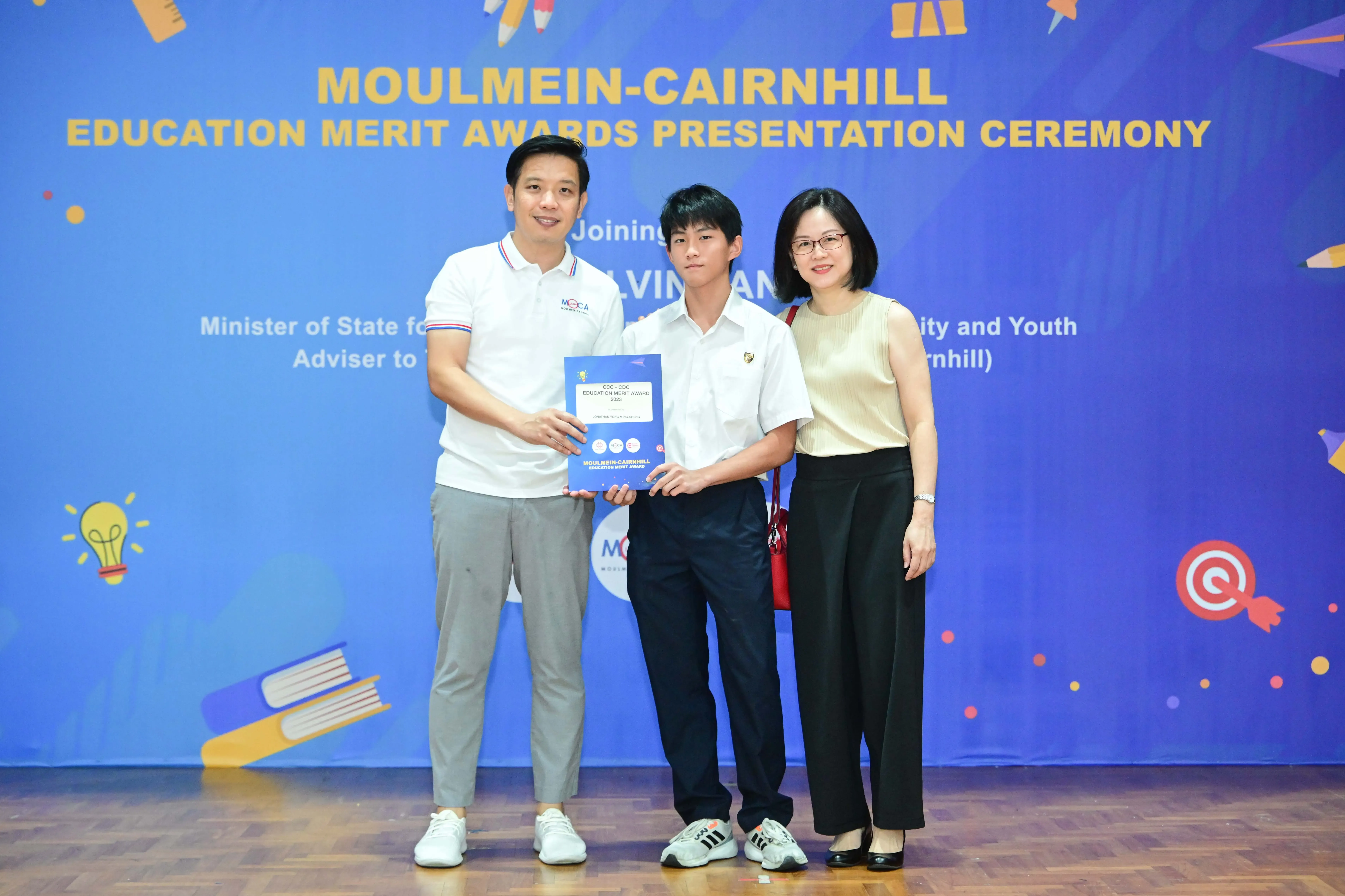 Three people stand before a Moulmein-Cairnhill Education Merit Awards banner, with a student holding a blue award certificate.