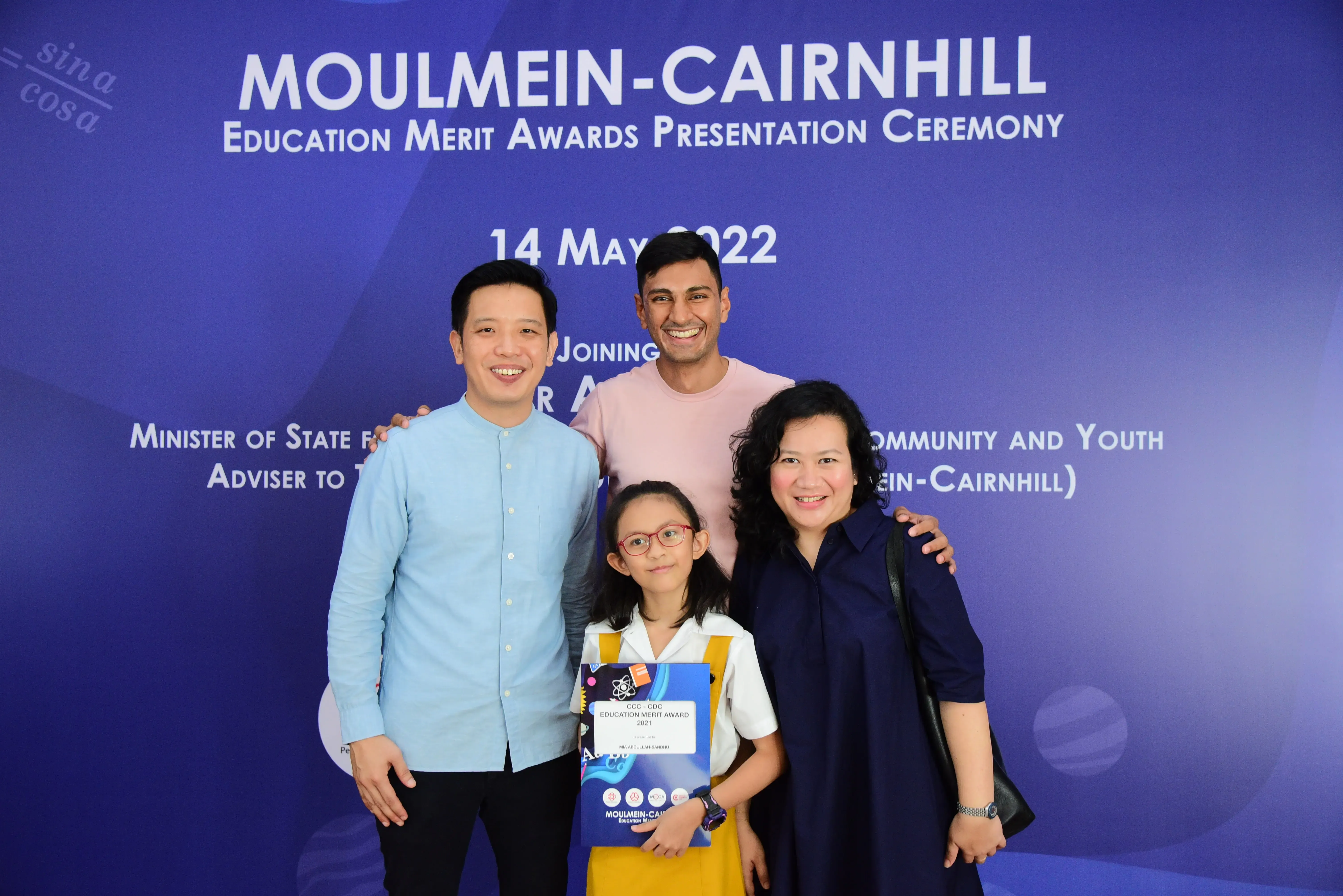 Three adults and a young girl holding an award certificate pose before a Moulmein-Cairnhill Education Merit Awards banner dated 14 May 2022.