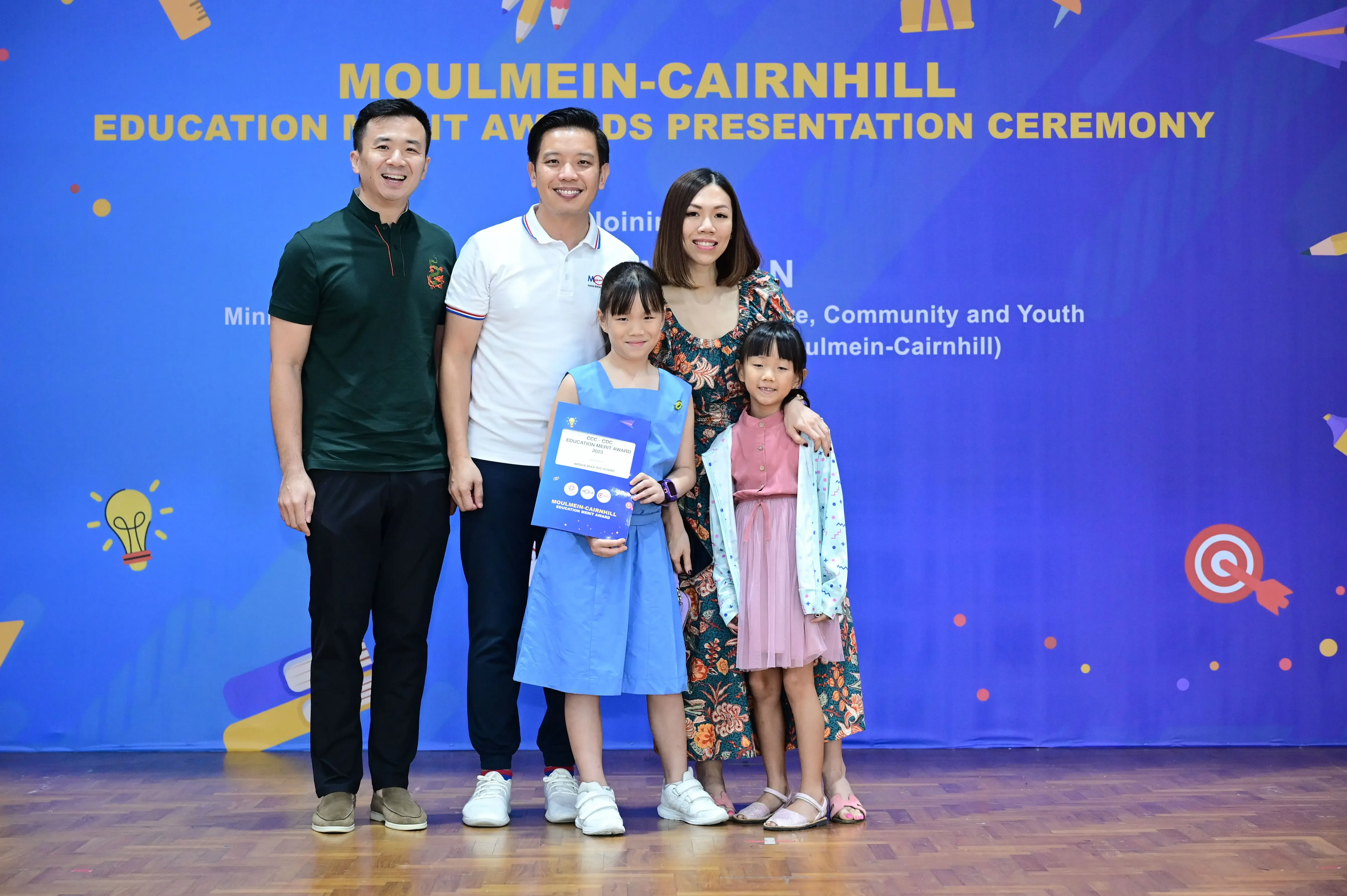 Five people pose at the Moulmein-Cairnhill Education Merit Awards Presentation Ceremony, with a girl holding an award certificate.