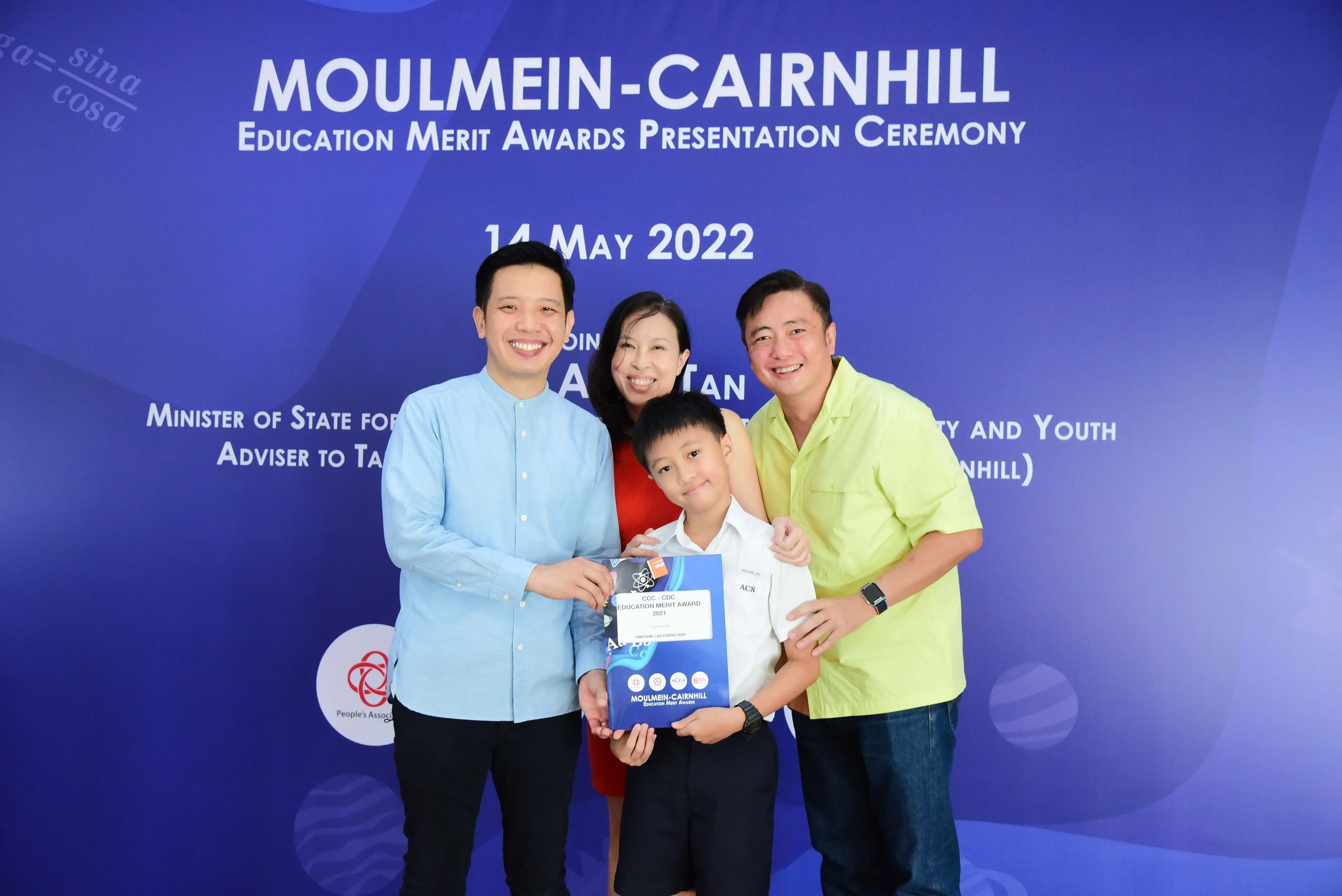 Four people smile before a Moulmein-Cairnhill Education Merit Awards backdrop dated 14 May 2022, with a boy holding an award.