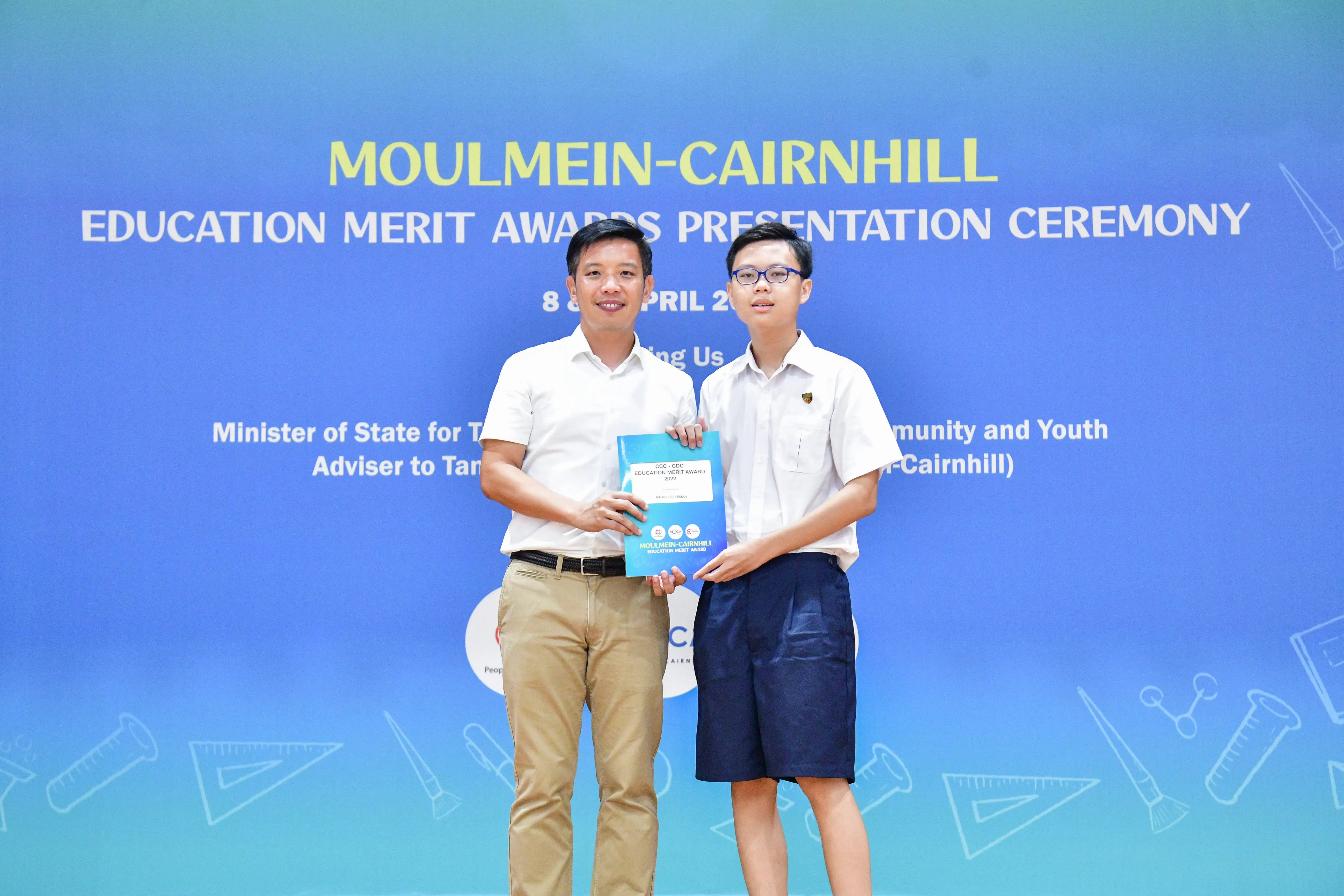 Two men hold a certificate together in front of a Moulmein-Cairnhill Education Merit Awards Presentation Ceremony banner.