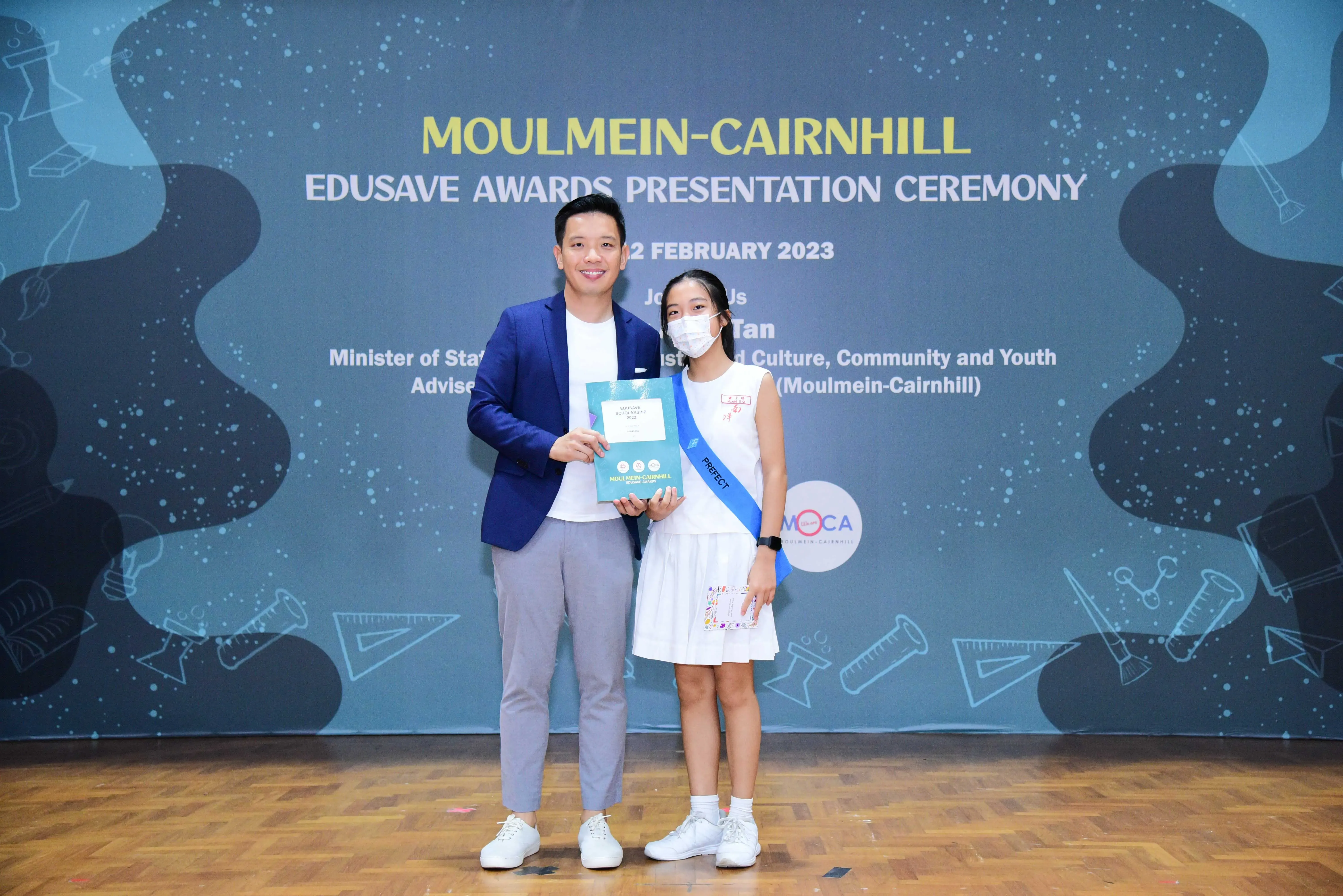 Man in blue blazer and masked girl wearing a Prefect sash pose holding an award at Moulmein-Cairnhill Edusave ceremony.