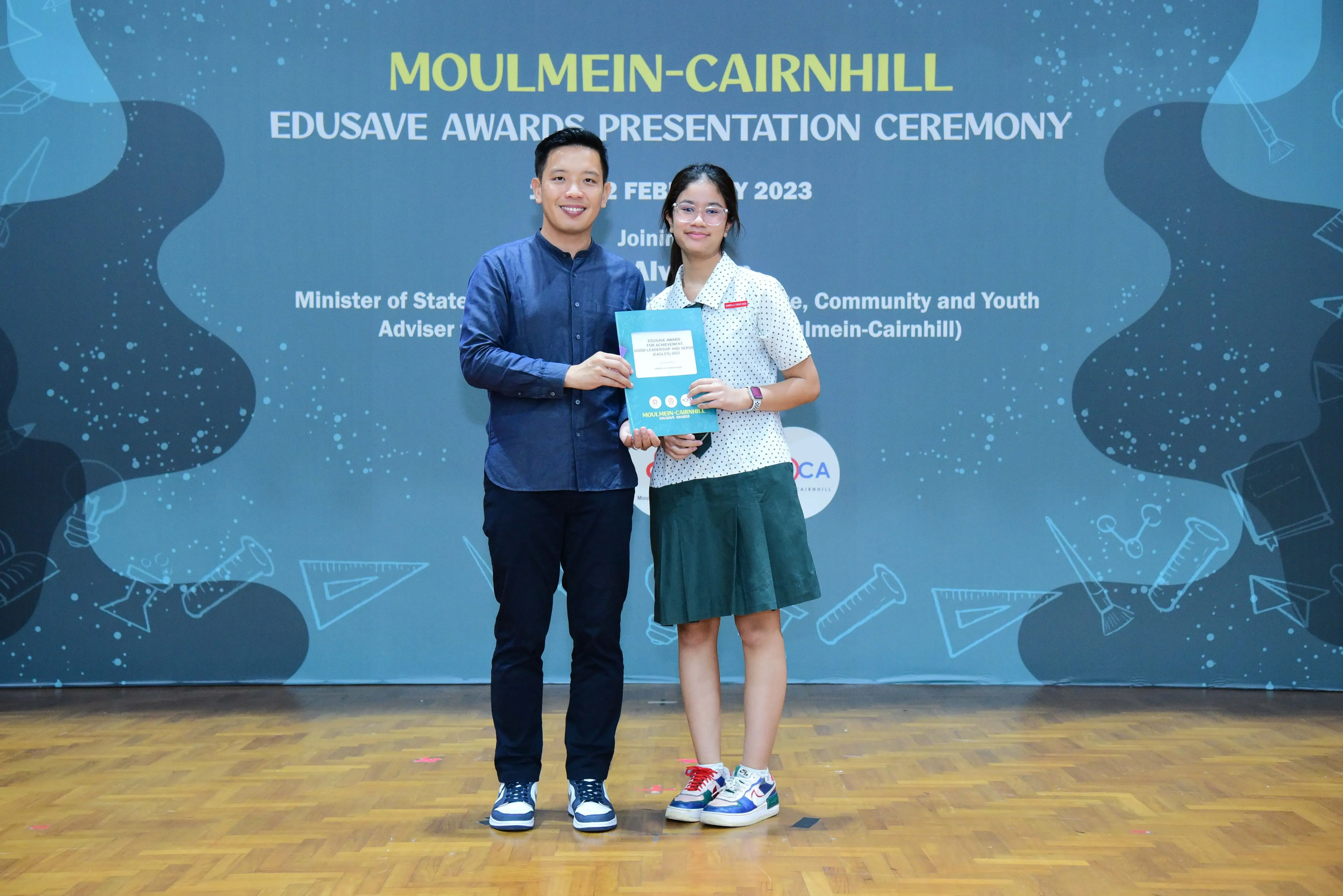 Two people hold a certificate together on stage at the Moulmein-Cairnhill Edusave Awards Presentation Ceremony 2023.