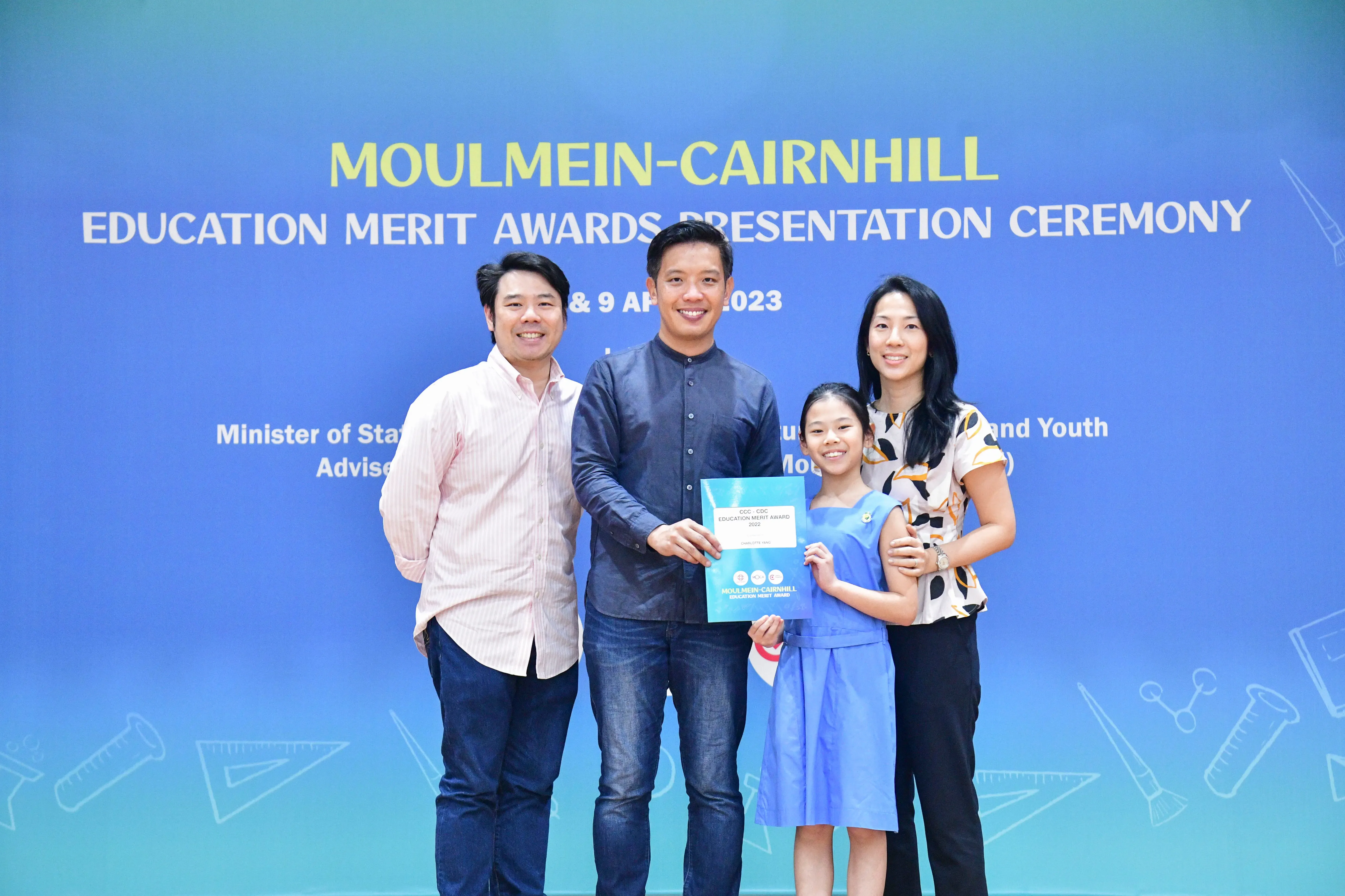 Four people pose before a Moulmein-Cairnhill Education Merit Awards 2023 banner, a girl holding a certificate.
