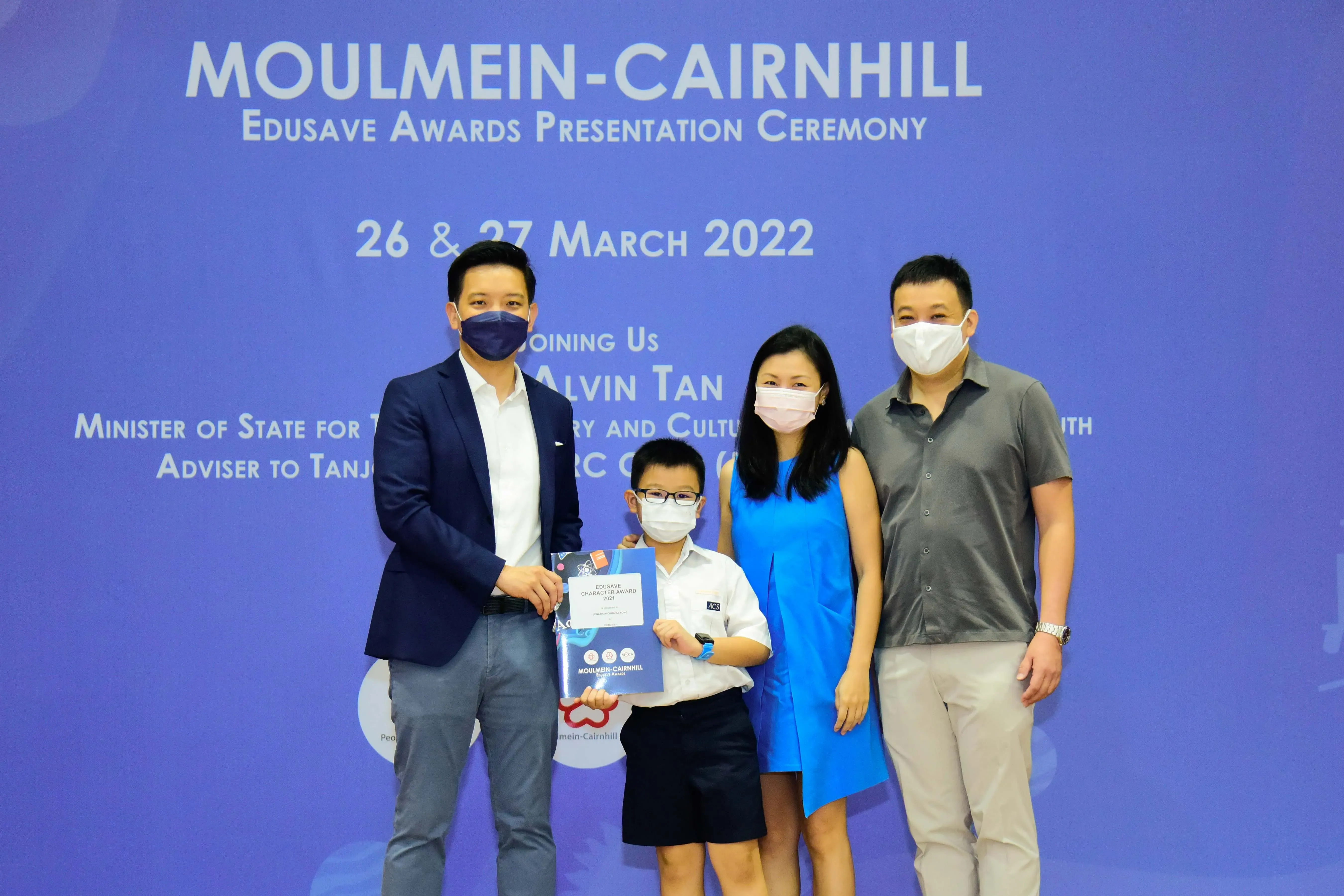 Four masked people pose at the Moulmein-Cairnhill Edusave Awards Ceremony on 26-27 March 2022, a boy holds a certificate.