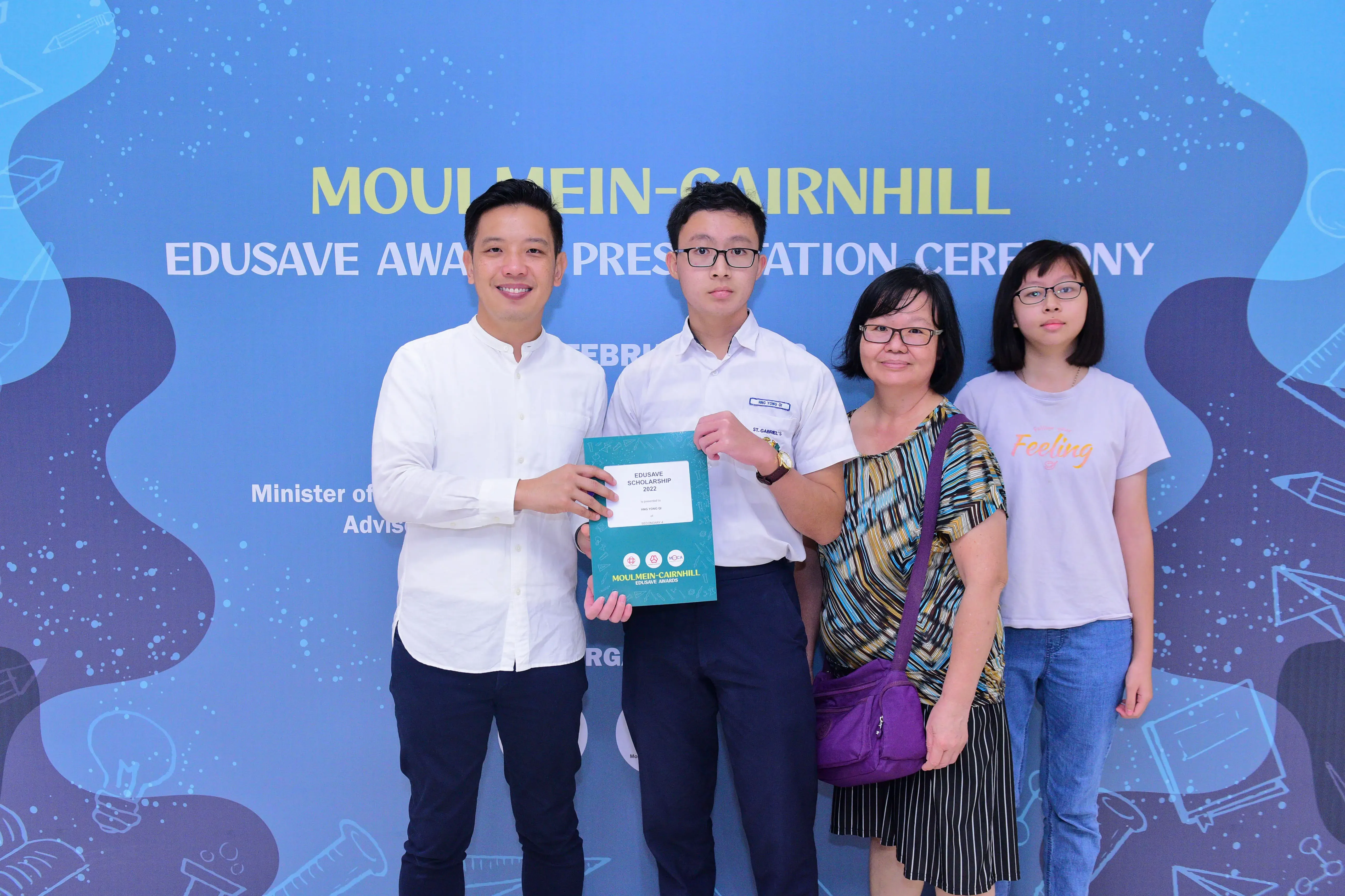 Four people pose at the Moulmein-Cairnhill Edusave Award Presentation Ceremony, holding a teal scholarship folder.