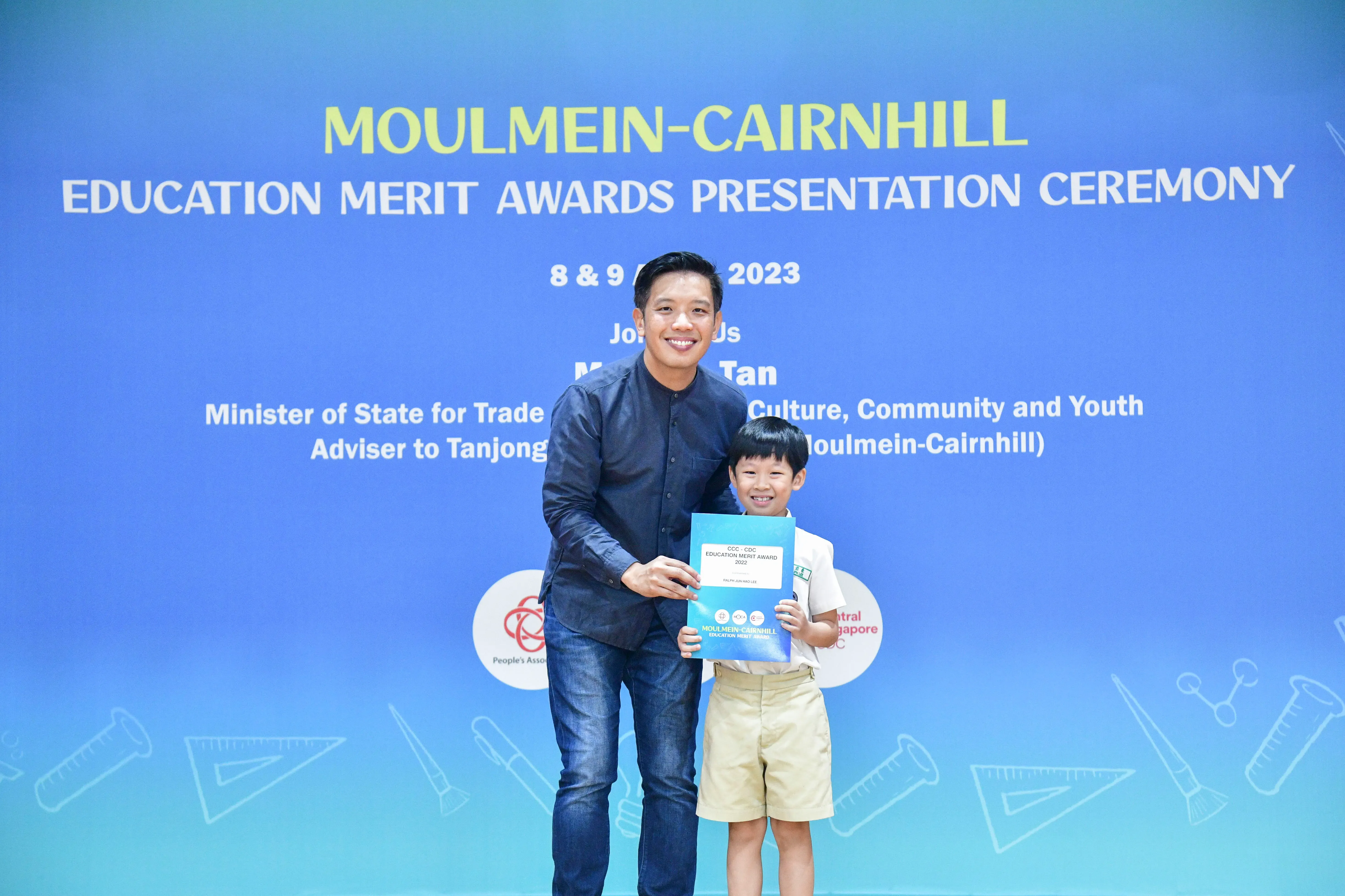 A man and a young boy hold a certificate together in front of a Moulmein-Cairnhill Education Merit Awards 2023 backdrop.