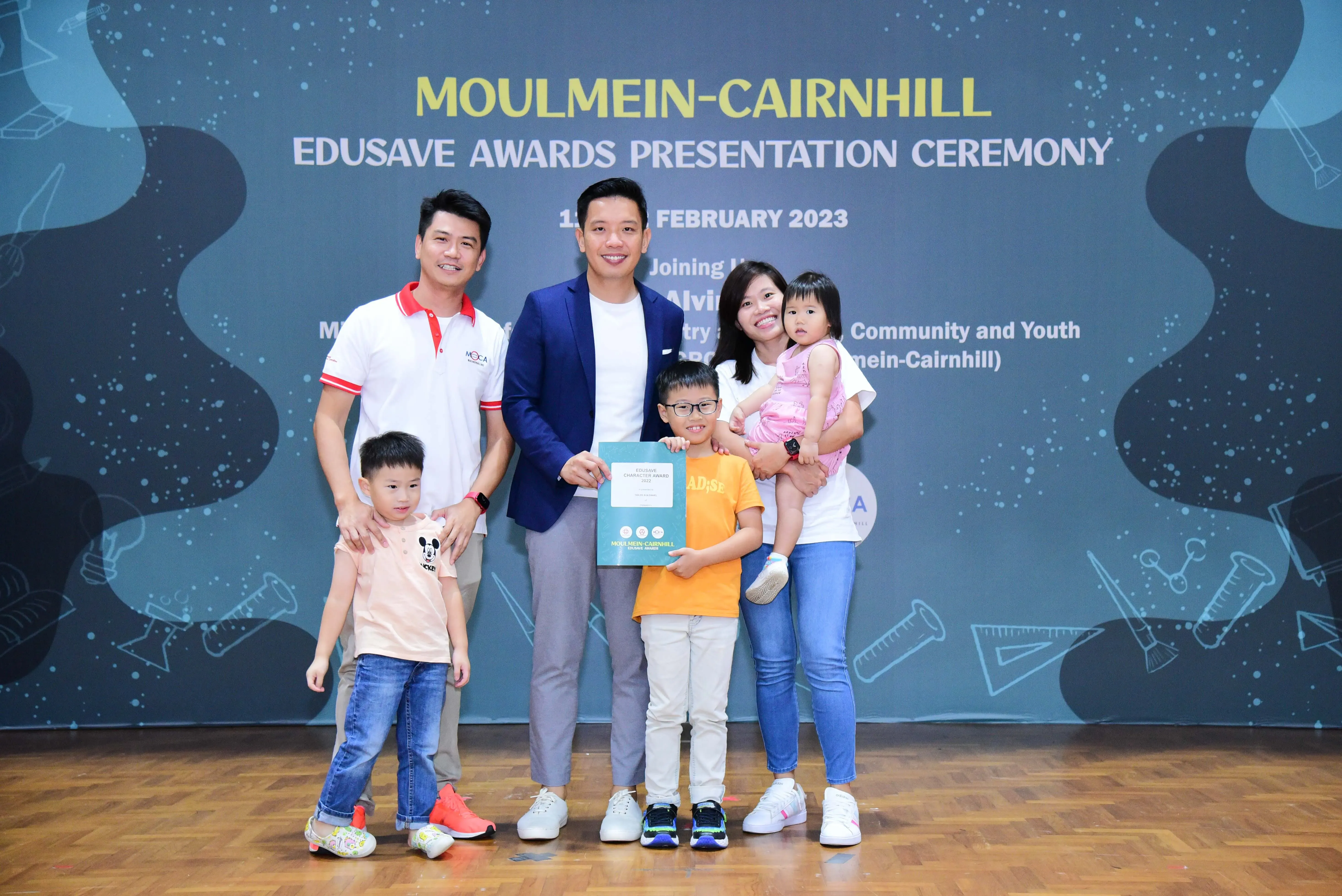 A family and a man in a blue blazer pose with a child holding a certificate at the Moulmein-Cairnhill Edusave Awards Ceremony.