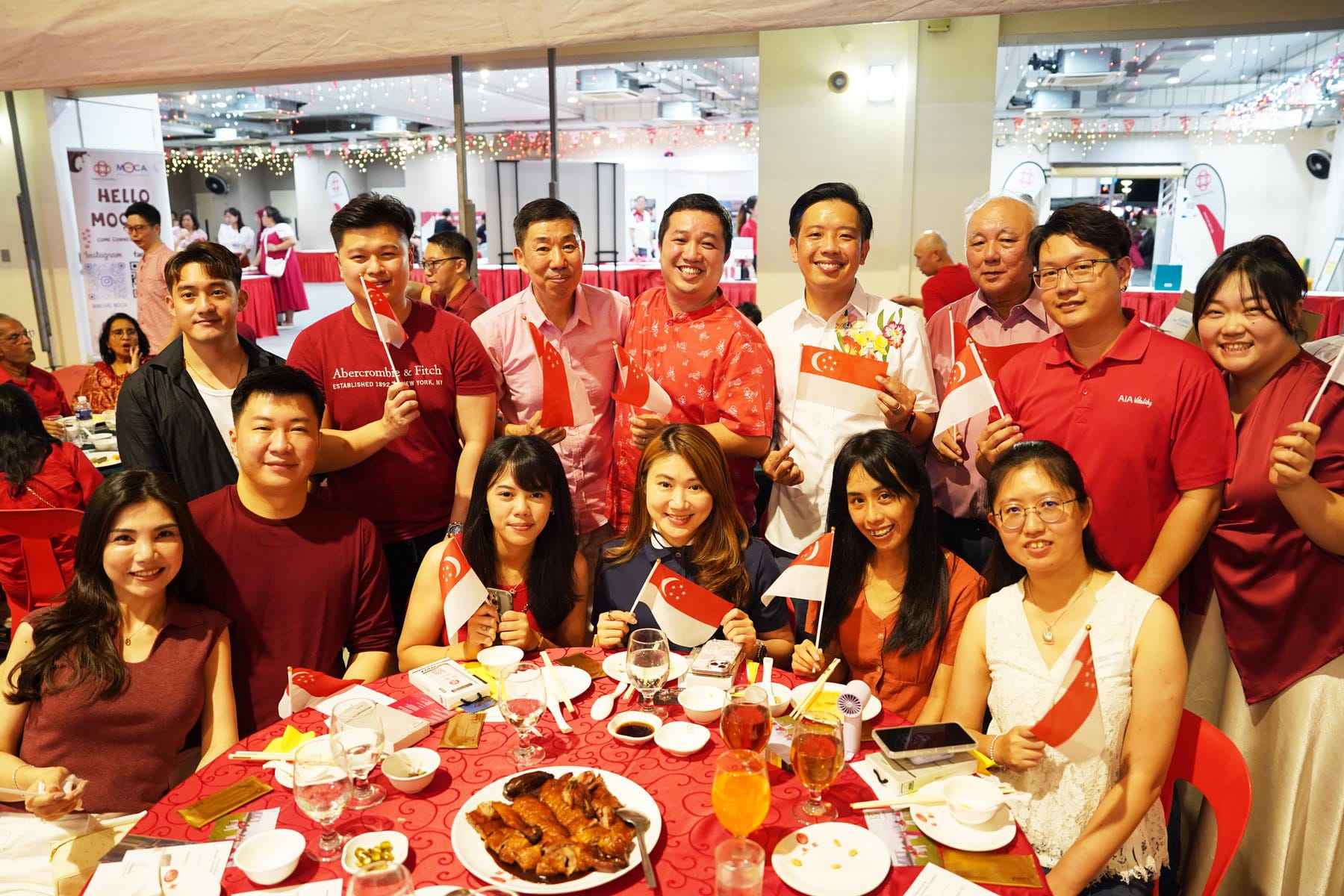 Group of people in red outfits smiling at a banquet table, holding small Singapore flags at a festive event.