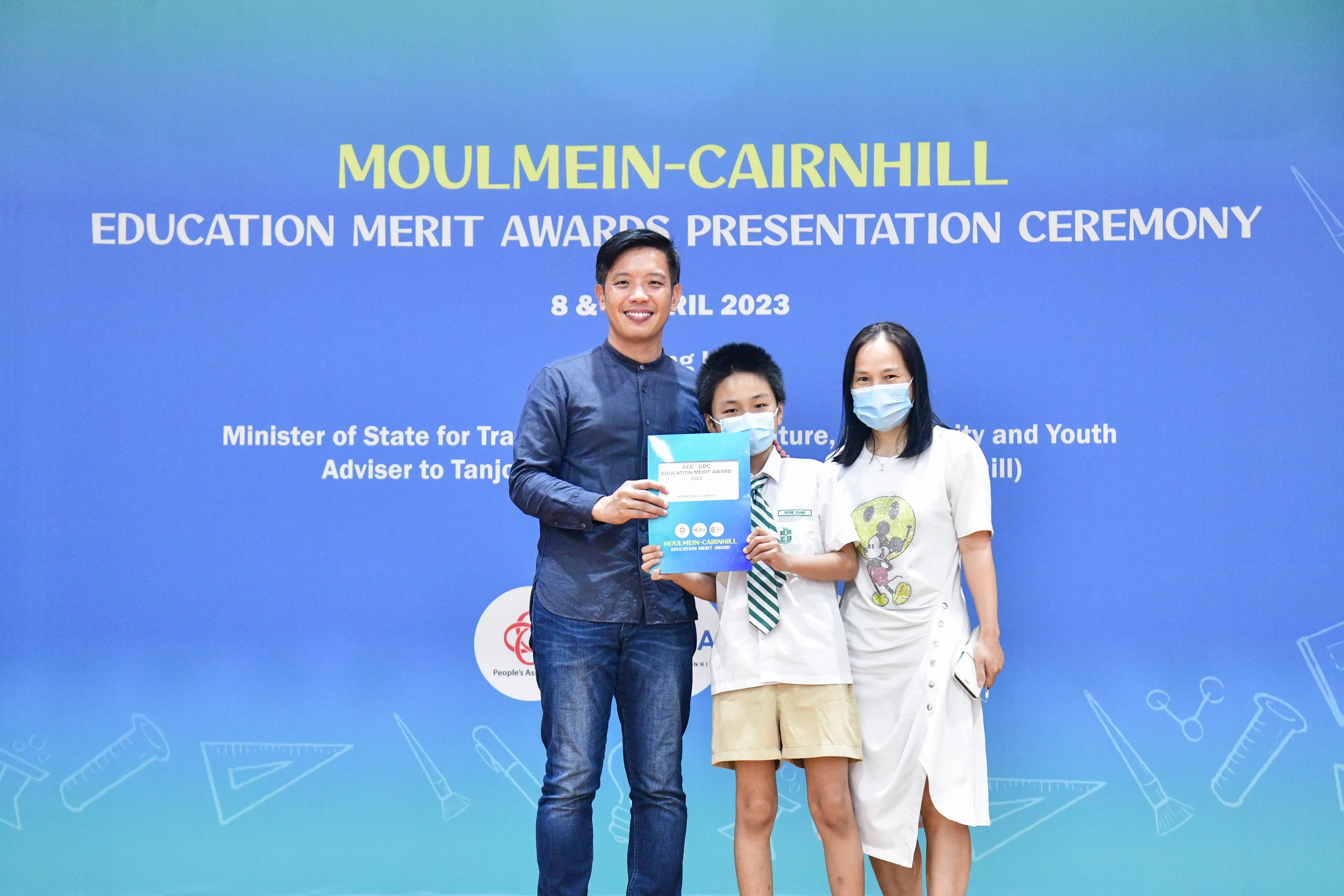 Three people pose holding an award certificate at the Moulmein-Cairnhill Education Merit Awards Ceremony, April 2023.