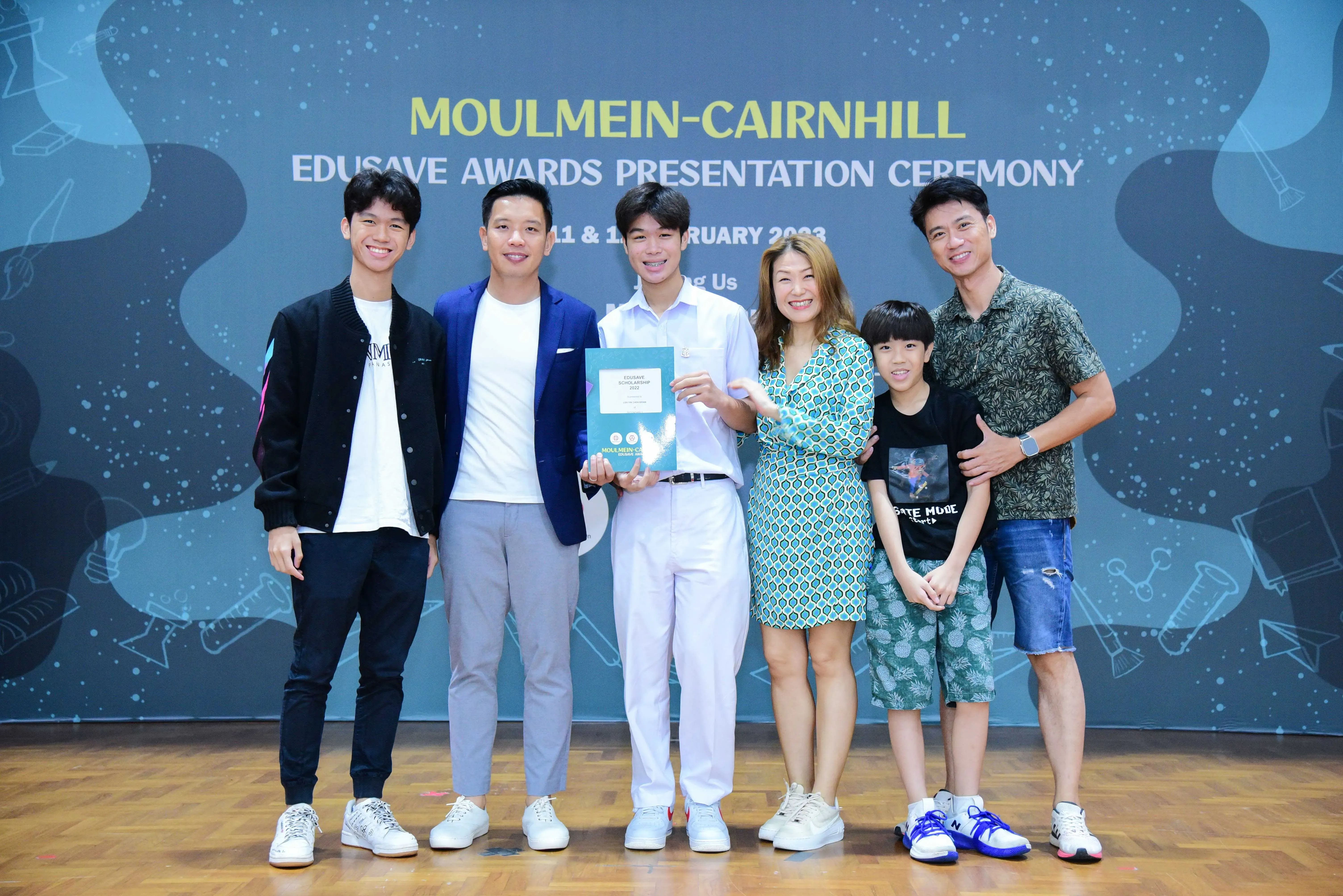 Six people smile while a young man holds an award certificate at the Moulmein-Cairnhill Edusave Awards Presentation Ceremony.