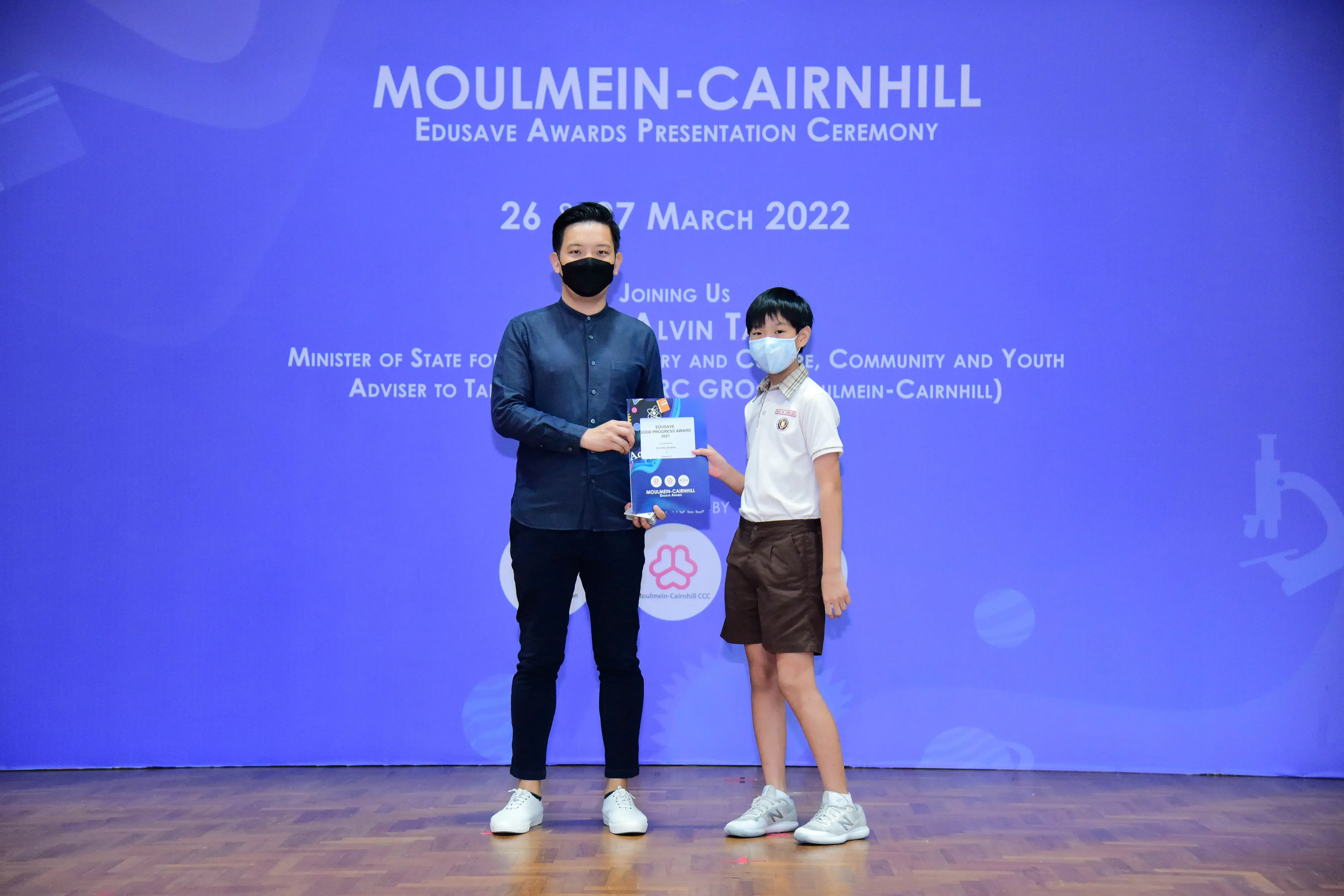 An adult and a young student hold an award together on stage at the Moulmein-Cairnhill Edusave Awards Ceremony 2022.