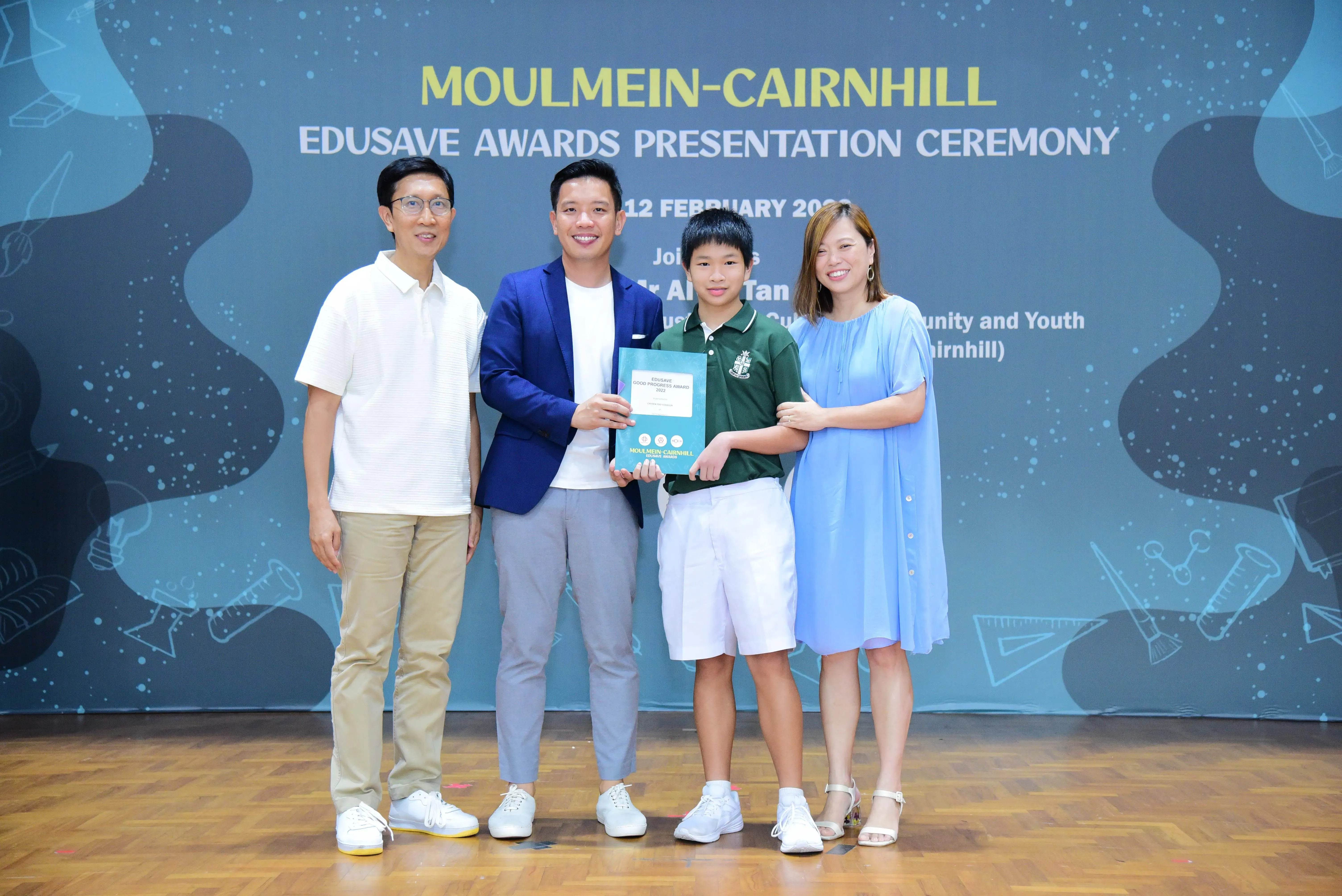 Four people pose at the Moulmein-Cairnhill Edusave Awards Presentation Ceremony, with a boy holding a certificate.