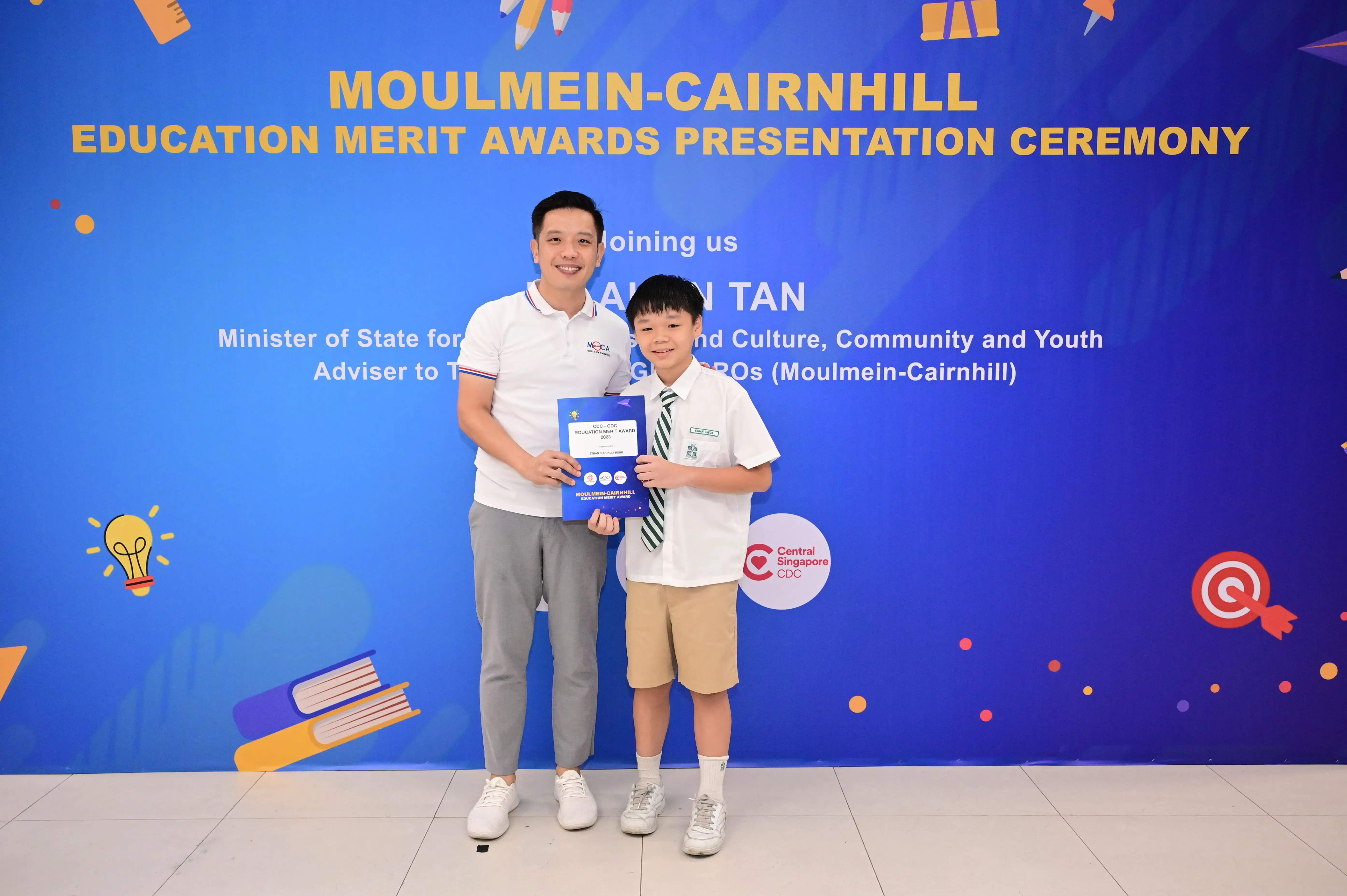 A man and a young student in uniform hold an award certificate in front of a Moulmein-Cairnhill Education Merit Awards banner.