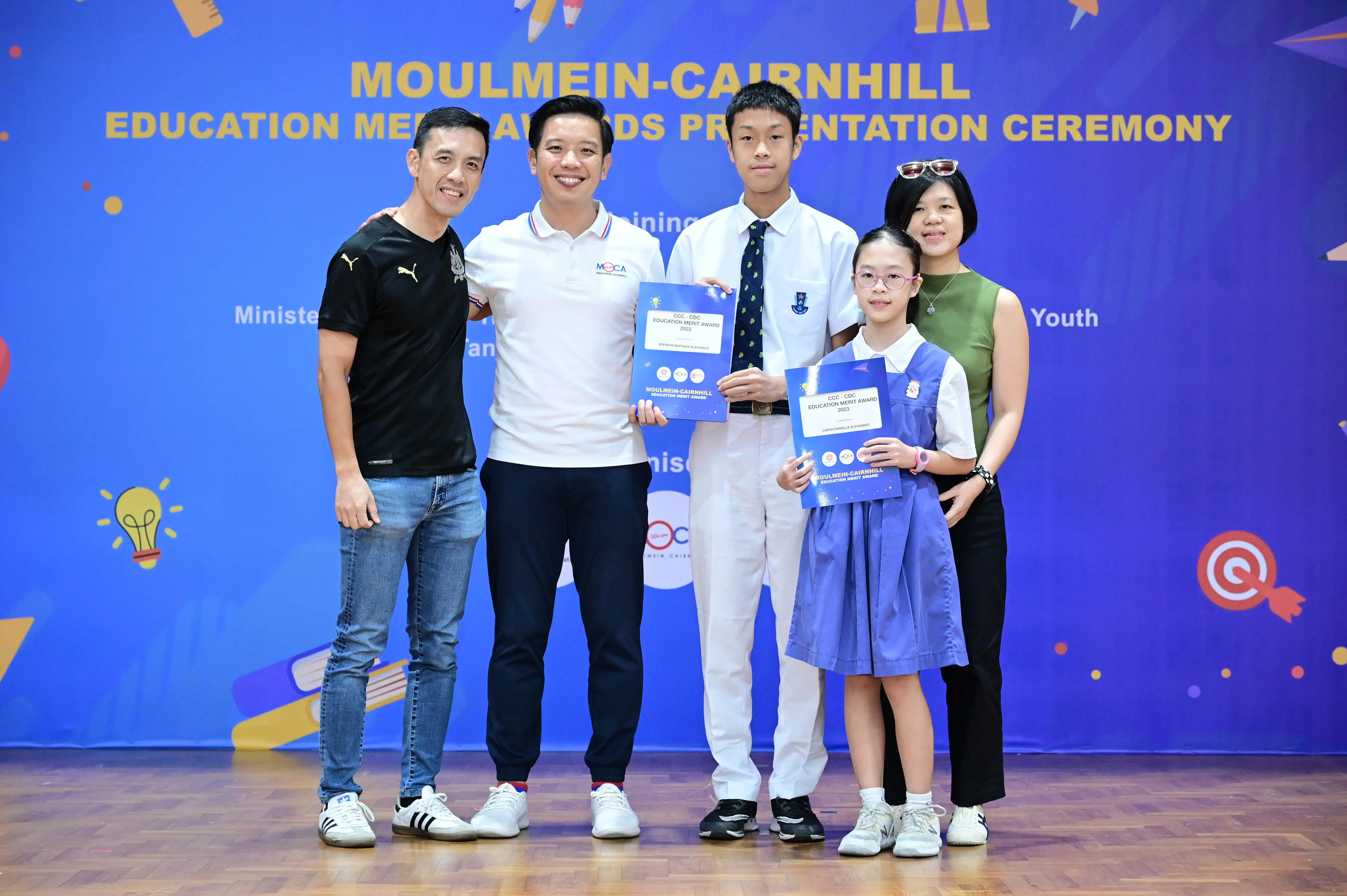 Five people pose at the Moulmein-Cairnhill Education Merit Awards Presentation Ceremony, two students holding award certificates.