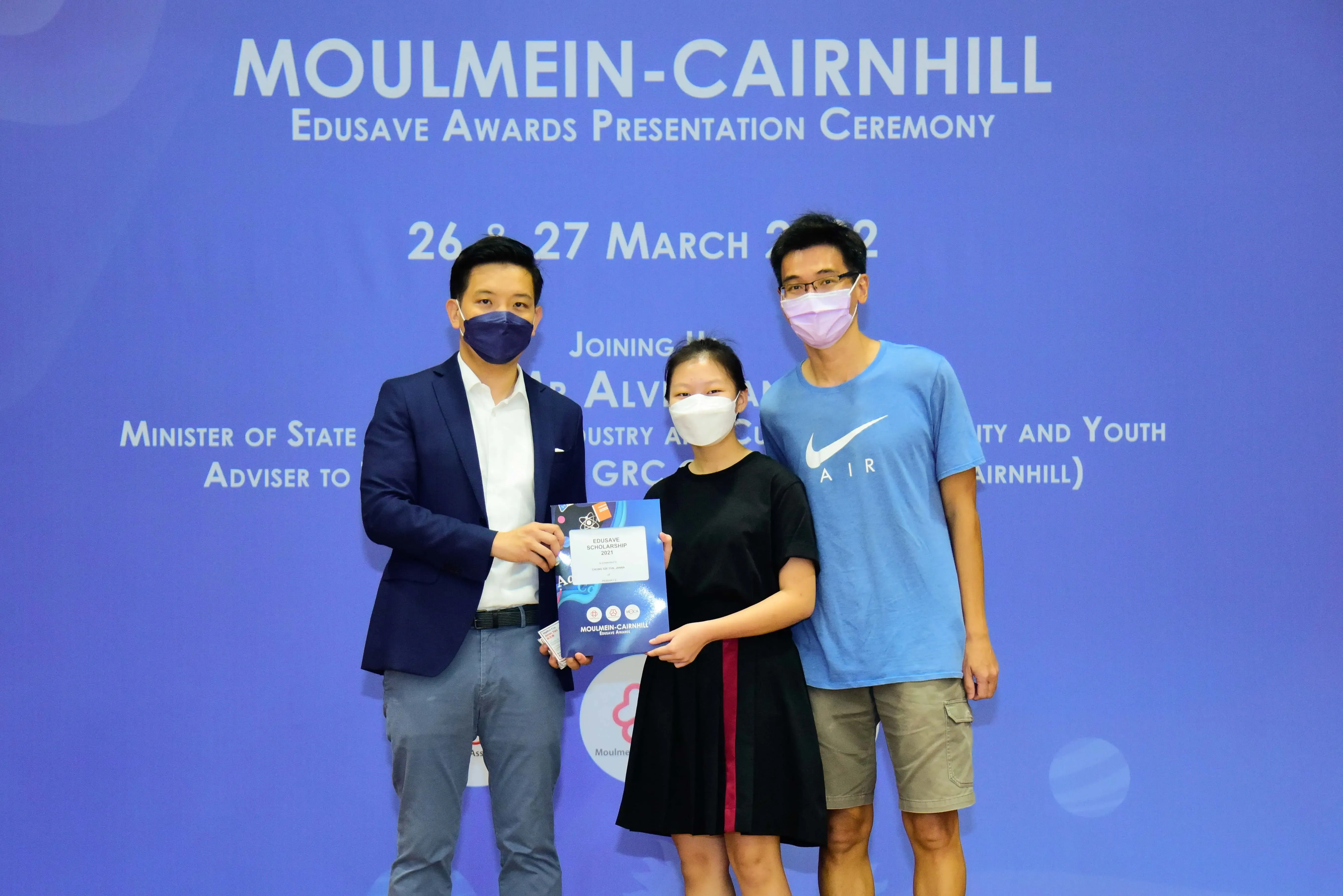 Three masked people pose at the Moulmein-Cairnhill Edusave Awards Presentation Ceremony, holding an award folder.
