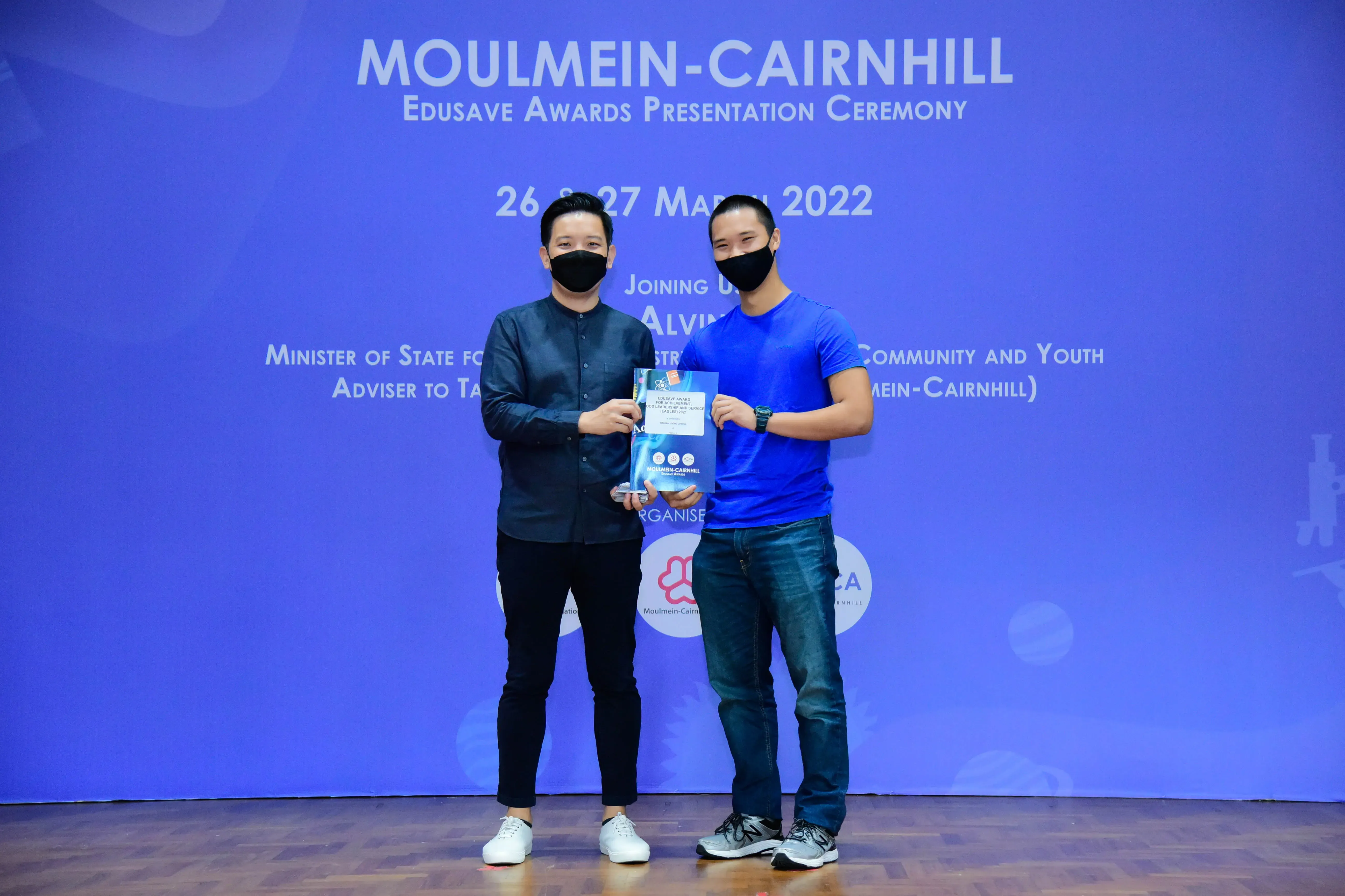 Two masked men hold an award booklet at the Moulmein-Cairnhill Edusave Awards Ceremony, March 2022.