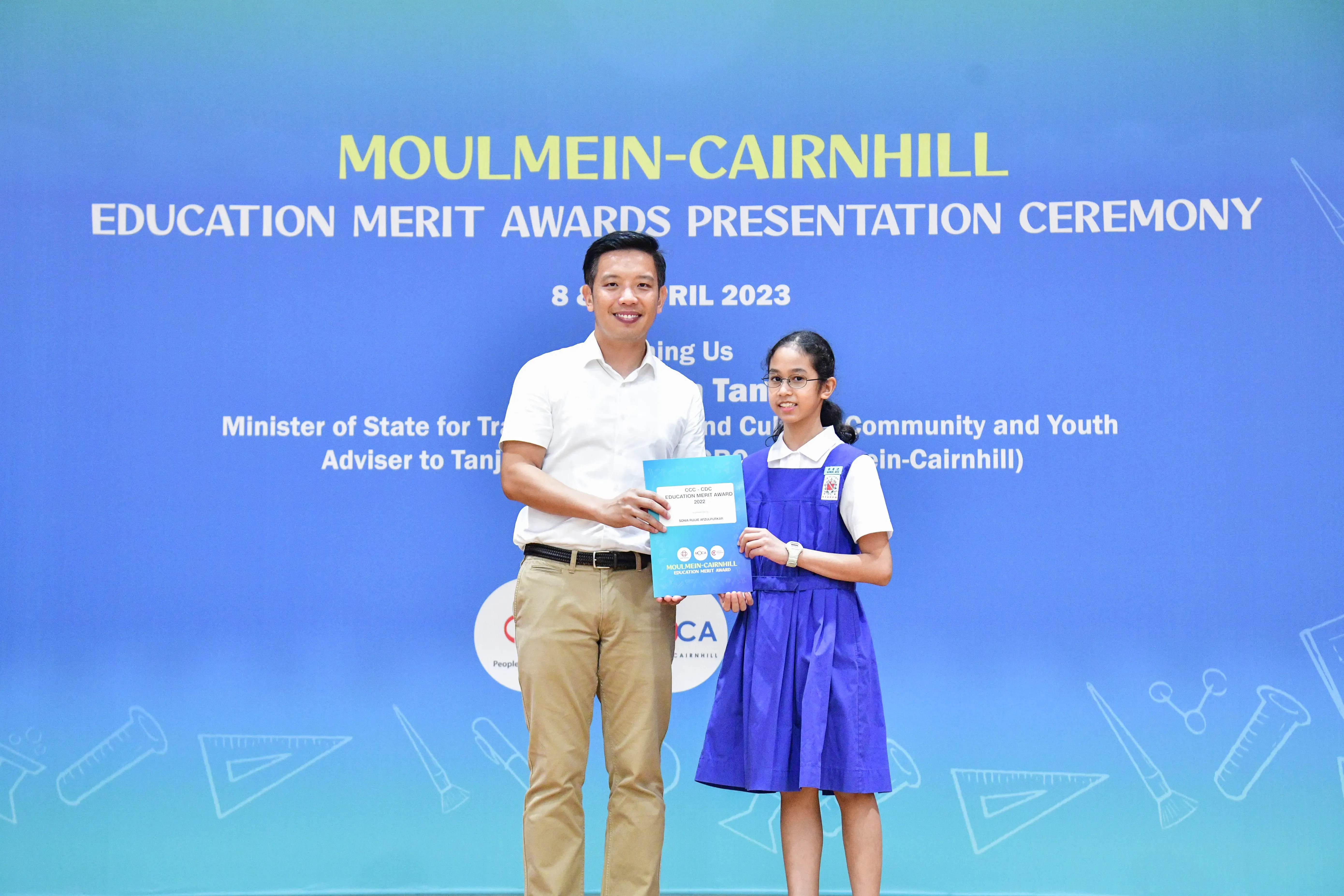 A man and a schoolgirl hold a blue award folder at the Moulmein-Cairnhill Education Merit Awards Ceremony, April 2023.