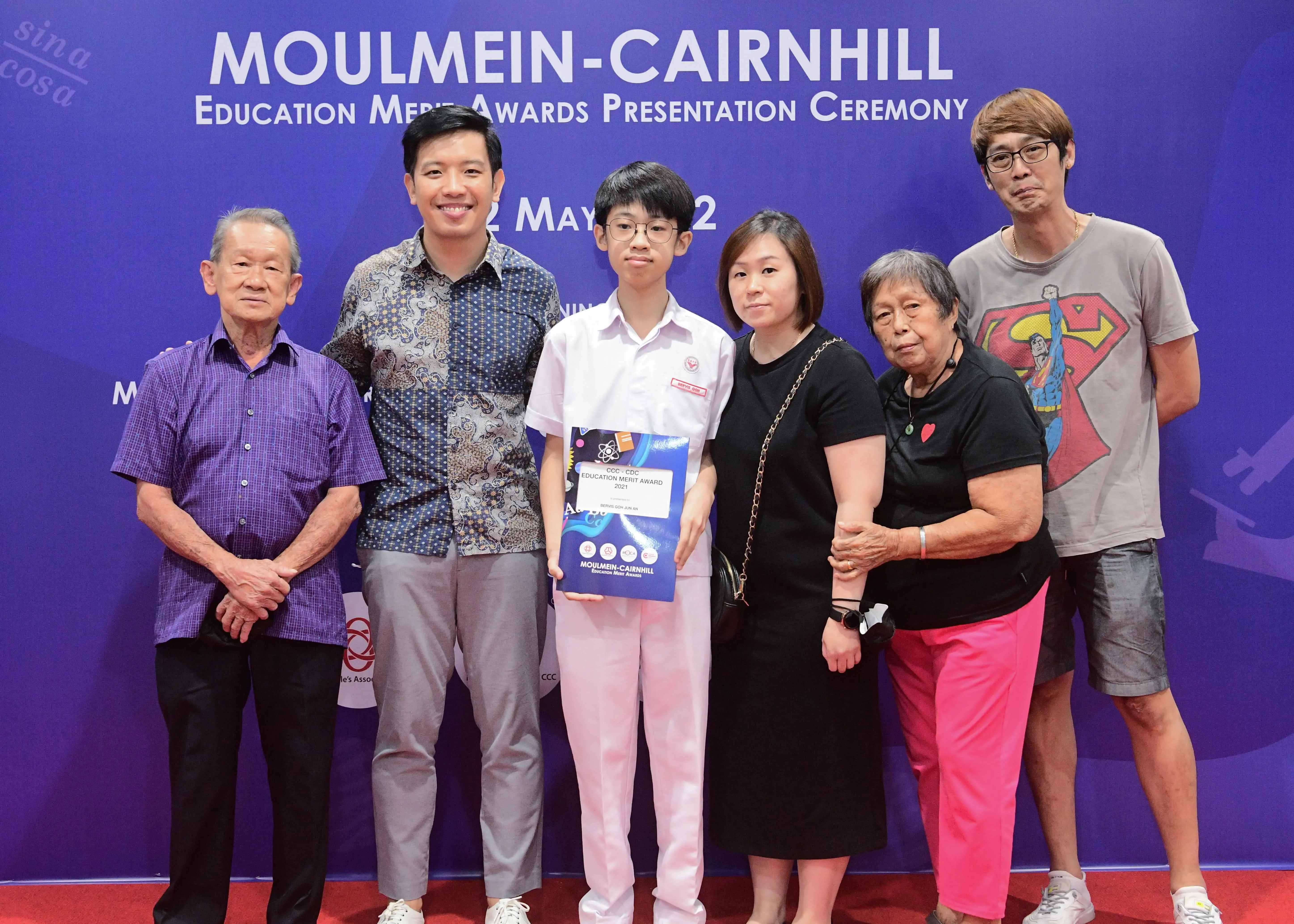 Six people pose in front of a Moulmein-Cairnhill Education Merit Awards banner, with a student holding an award certificate.