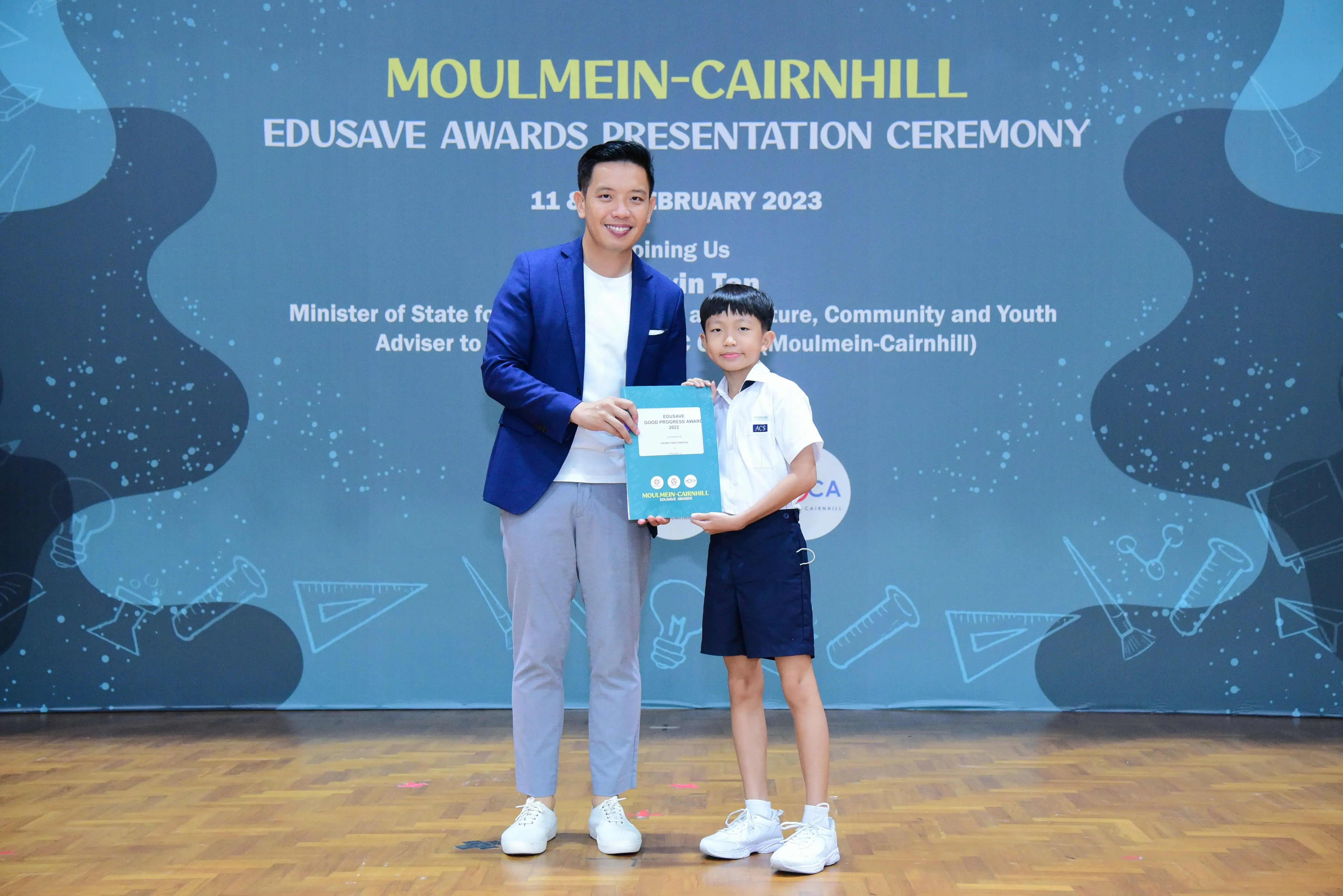 A man in a blue blazer and a young boy in school uniform hold an award at the Moulmein-Cairnhill Edusave Awards Ceremony 2023.