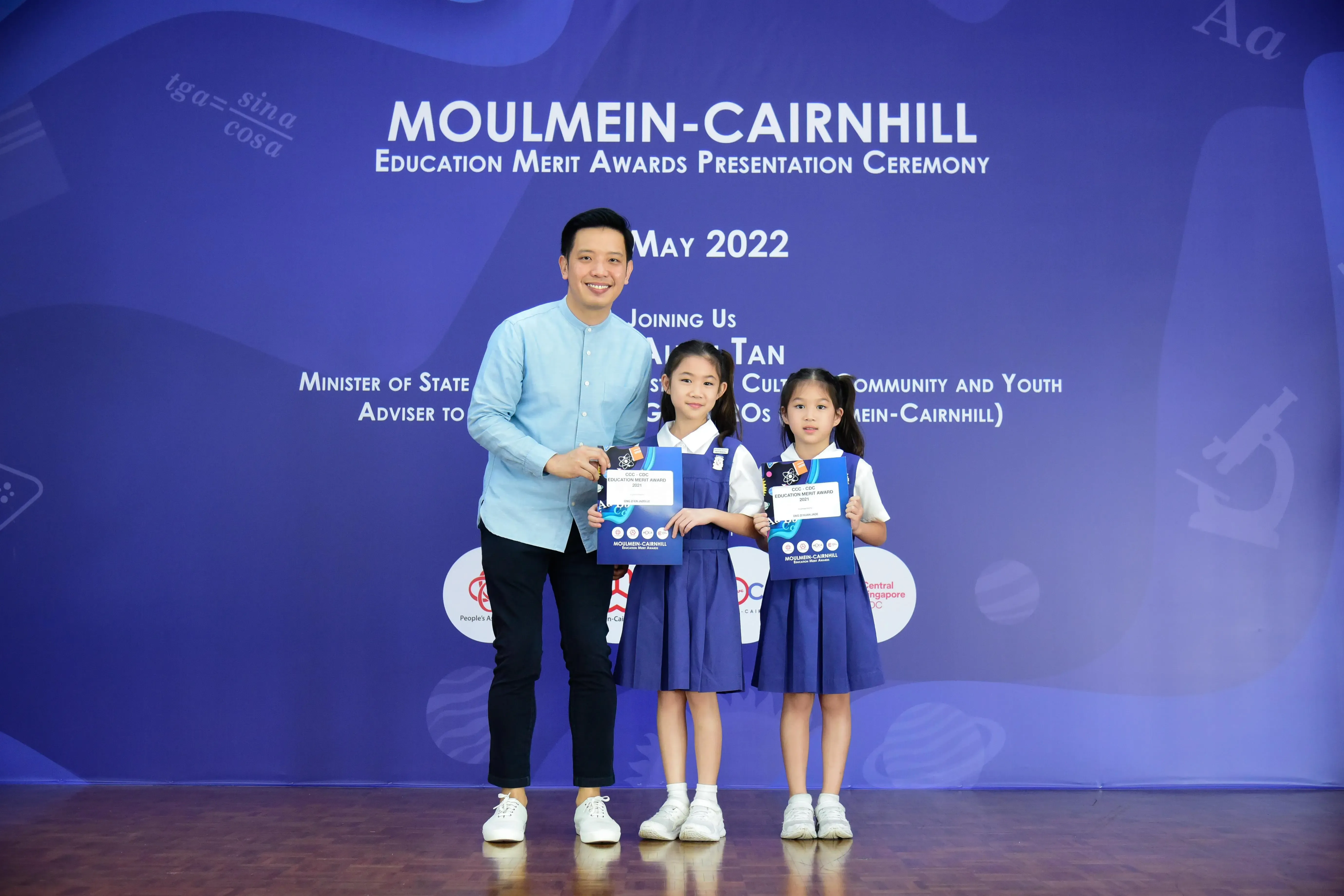 A man poses with two young girls holding award folders at the Moulmein-Cairnhill Education Merit Awards May 2022.