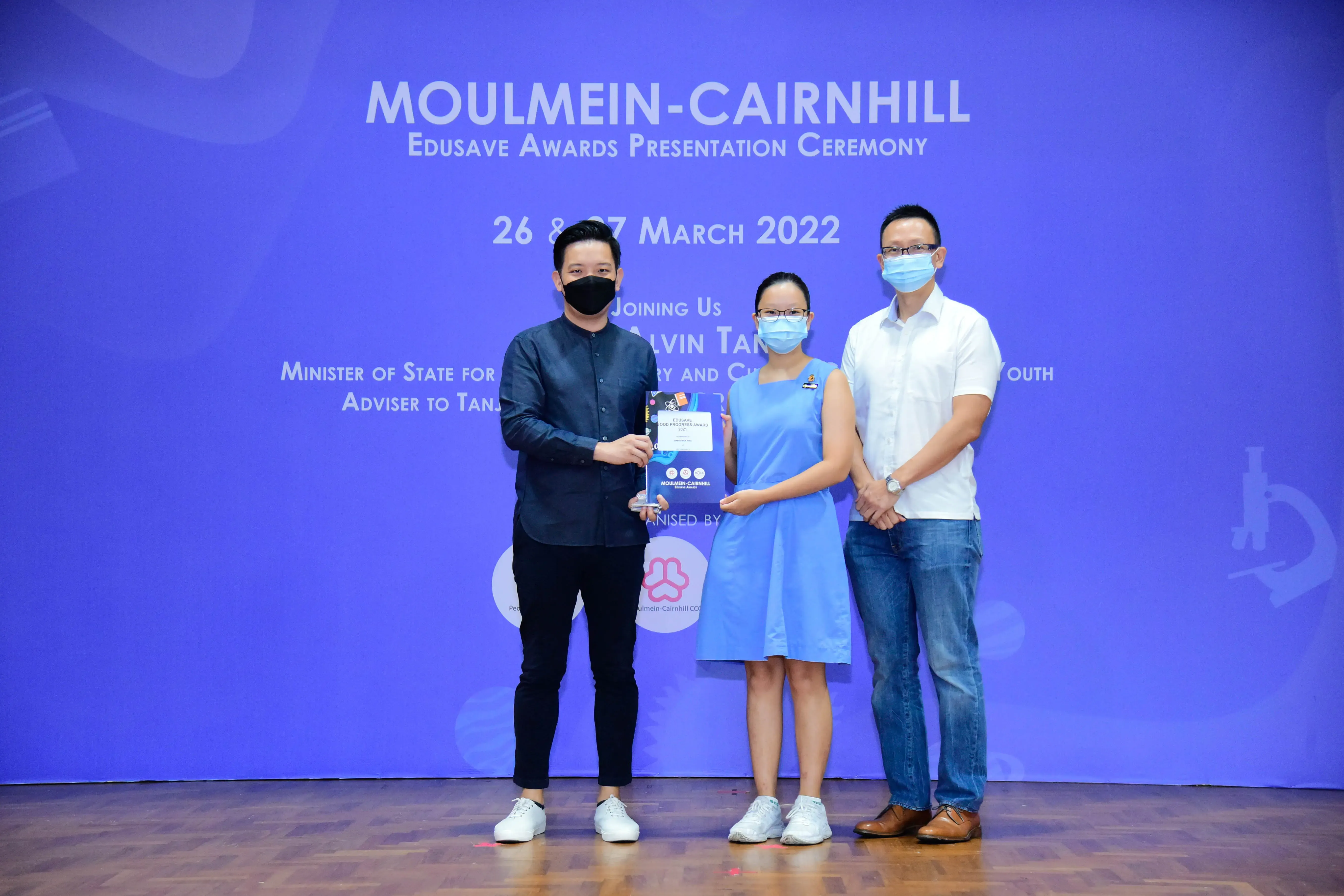 Three masked people stand on a stage holding an award at the Moulmein-Cairnhill Edusave Awards Ceremony, March 2022.