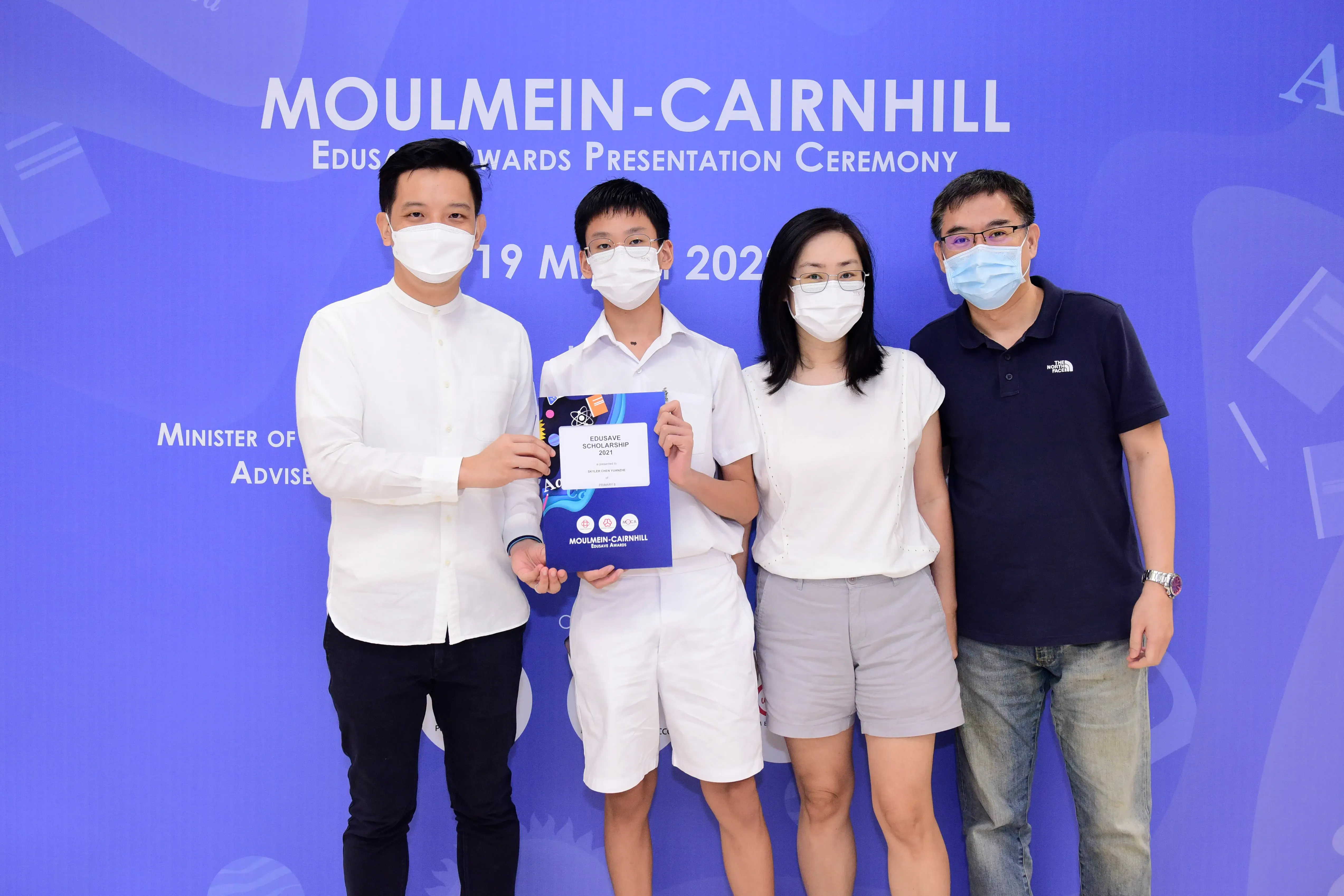 Four masked people pose at the Moulmein-Cairnhill Edusave Awards Presentation Ceremony, with a boy holding a certificate.