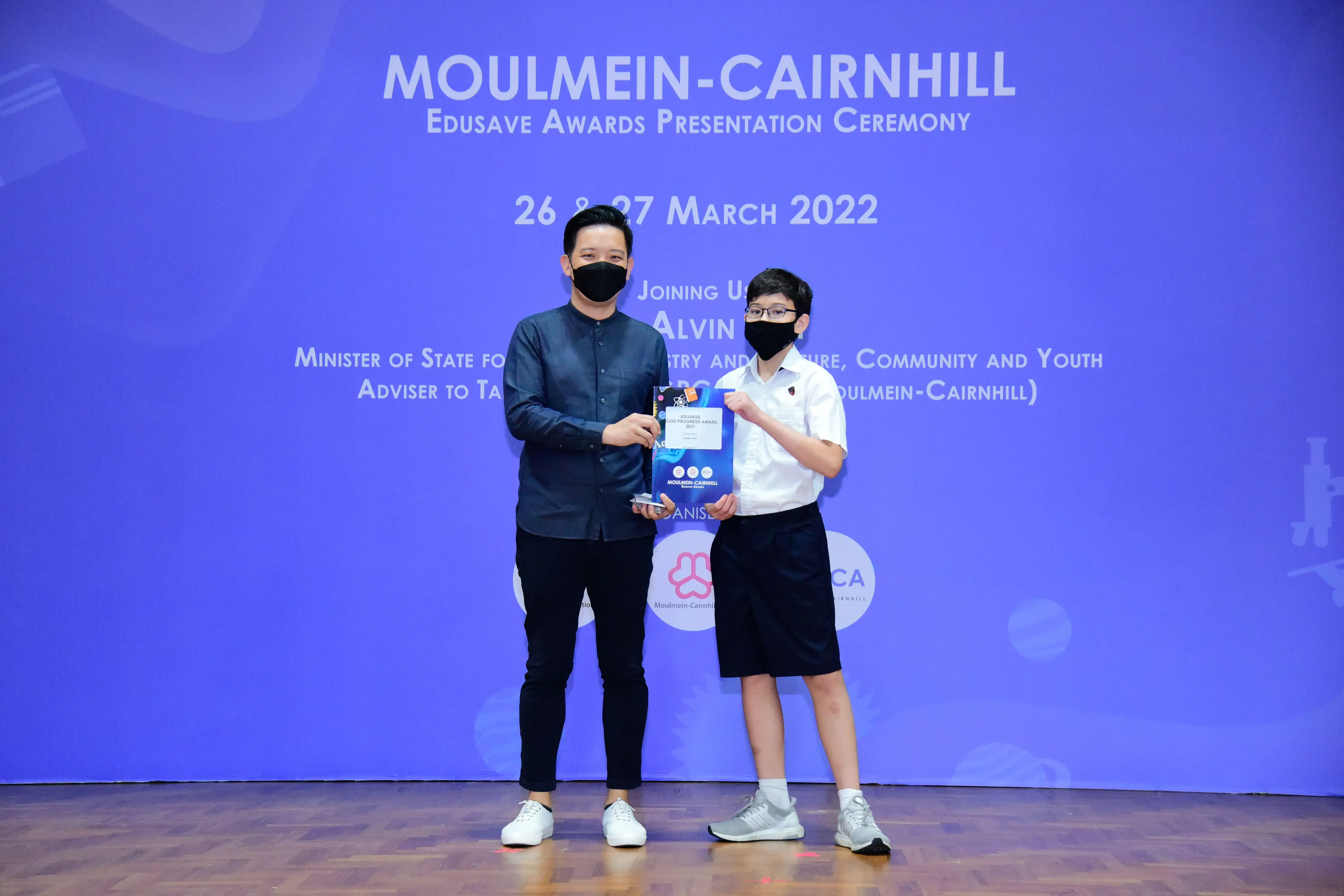 An adult and a young student in masks hold an award at the Moulmein-Cairnhill Edusave Awards Ceremony, March 2022.