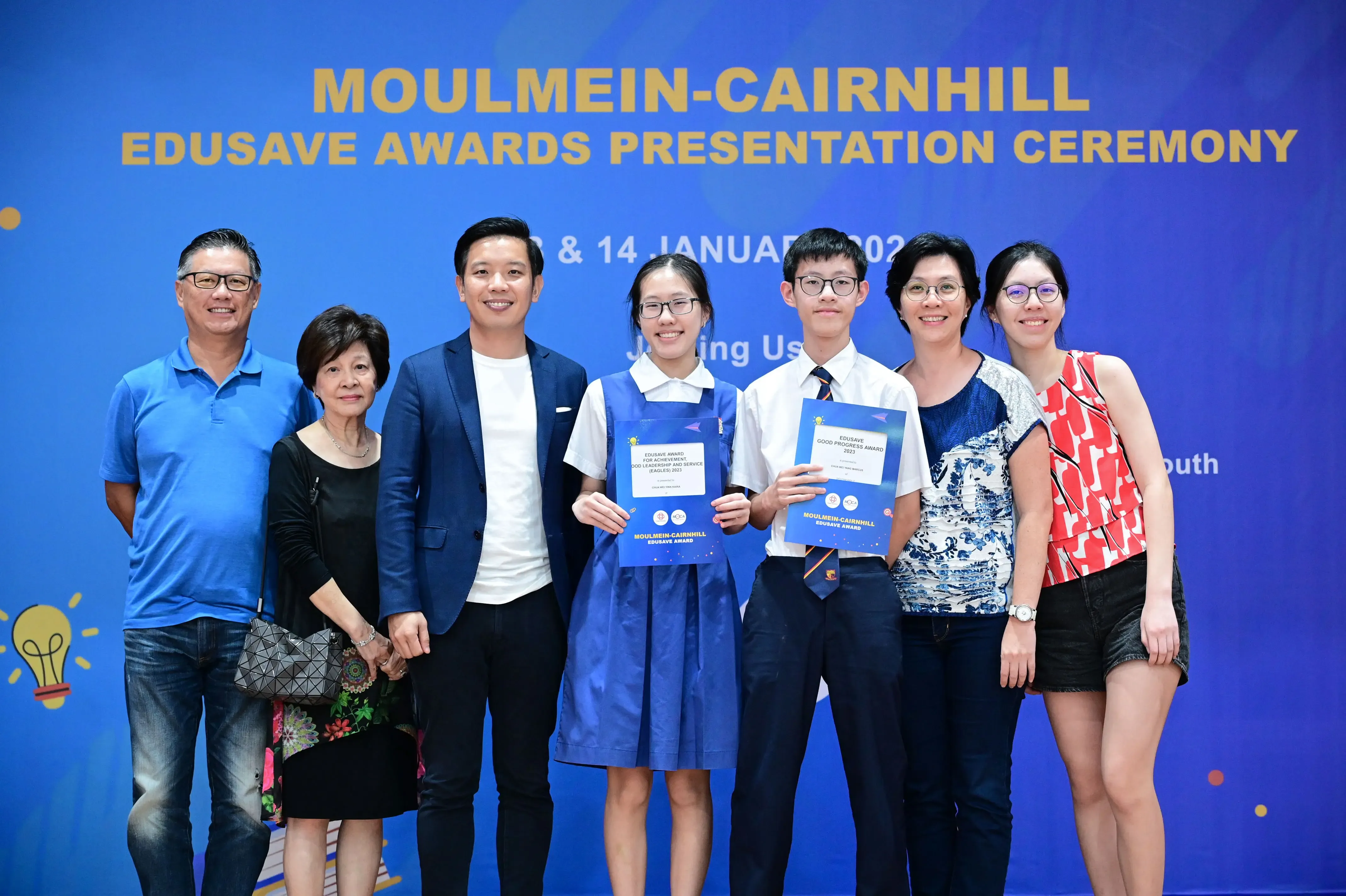Seven people pose at the Moulmein-Cairnhill Edusave Awards Presentation Ceremony, two students holding award certificates.