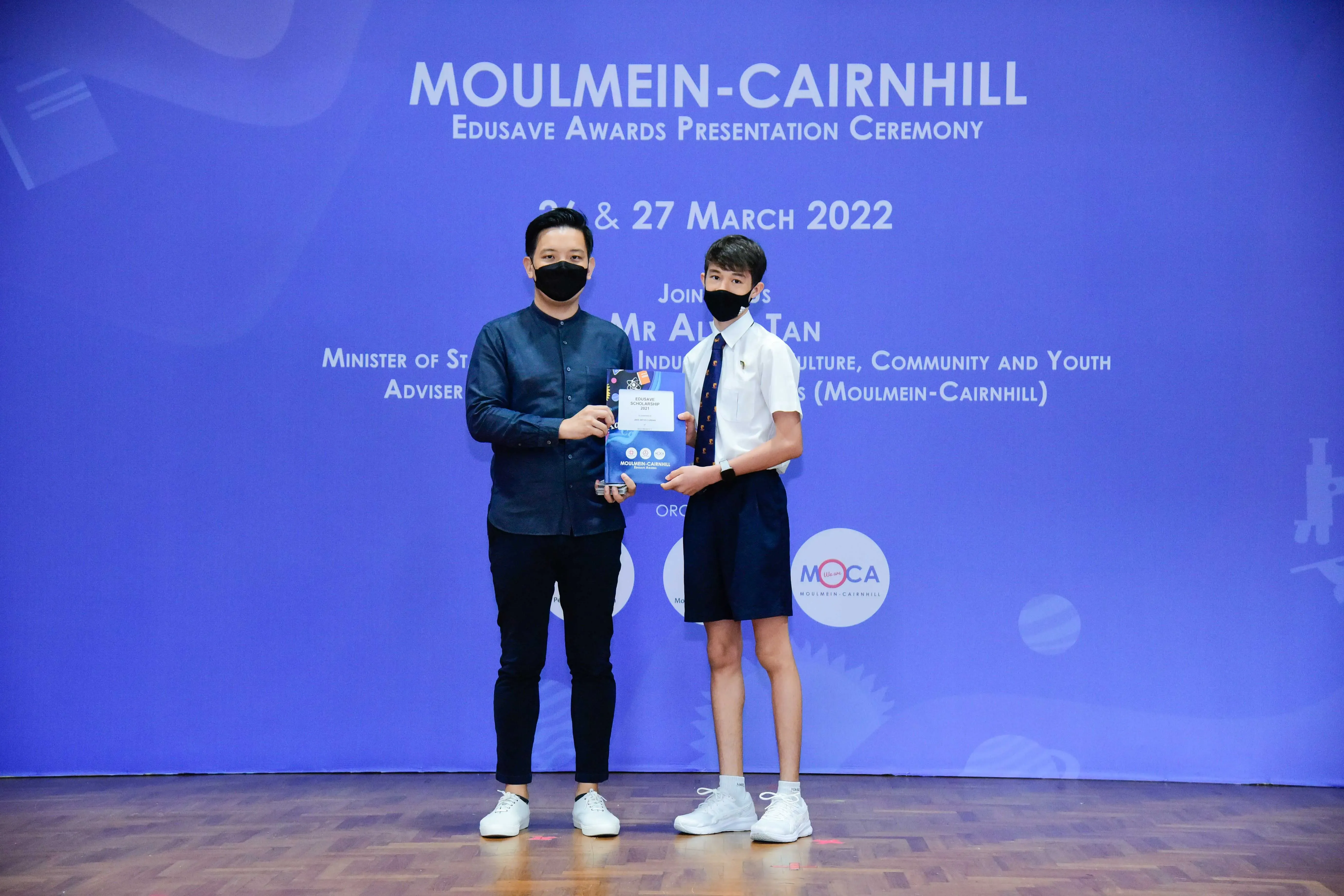 Two masked people hold an award at the Moulmein-Cairnhill Edusave Awards Presentation Ceremony, March 2022.
