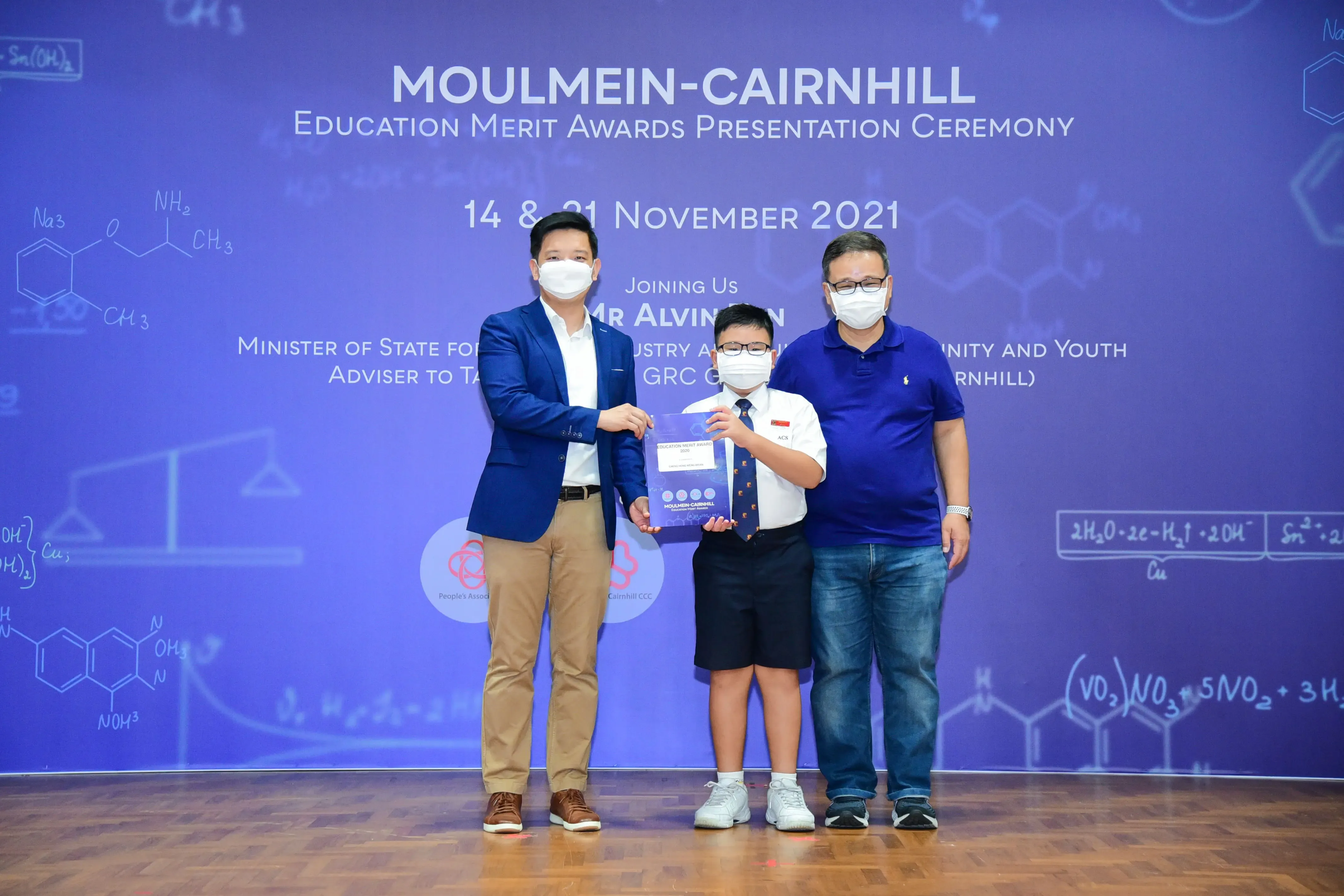 Two masked adults and a masked child hold an award at the Moulmein-Cairnhill Education Merit Awards Ceremony, November 2021.