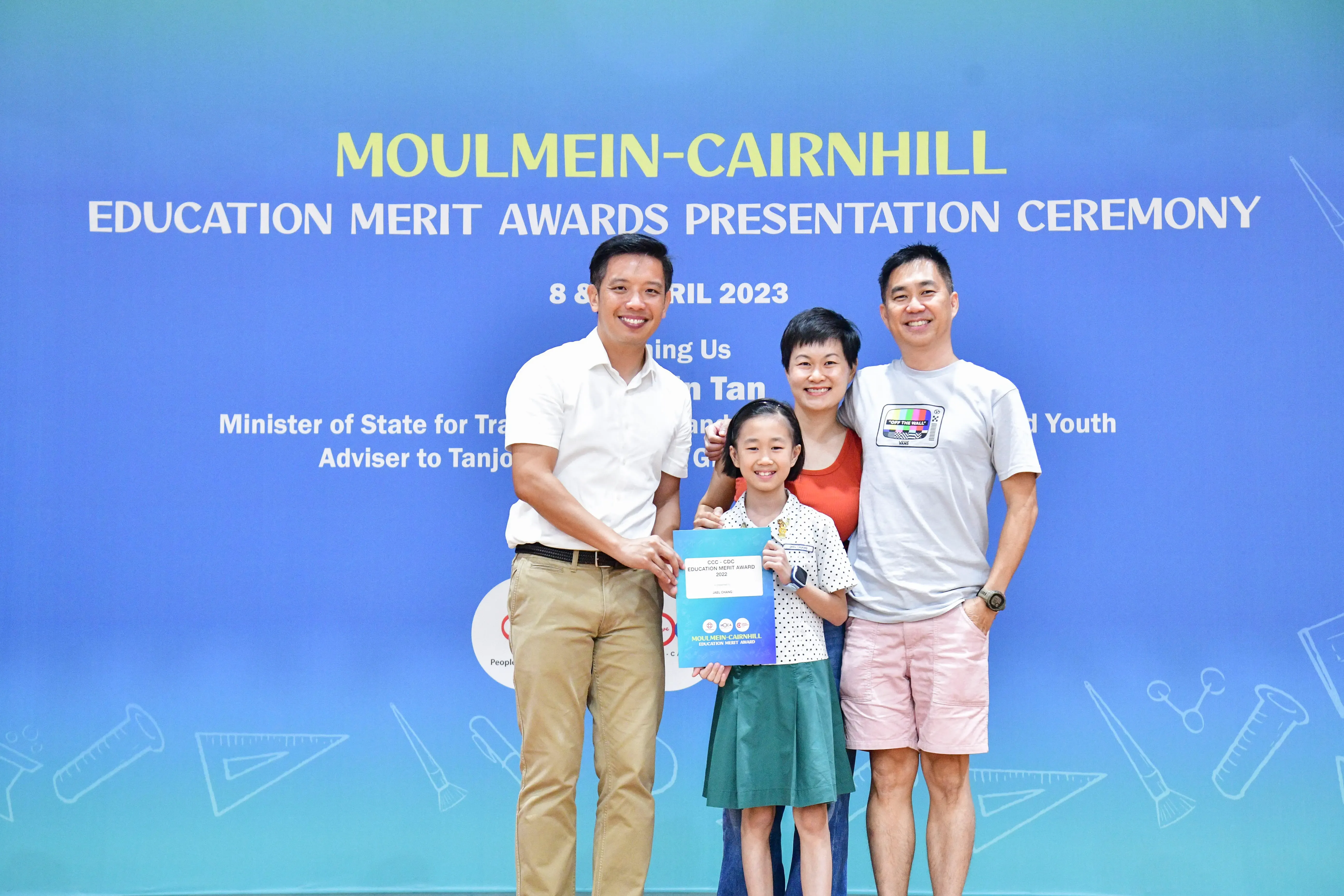 Four people smile before a Moulmein-Cairnhill Education Merit Awards banner; a girl holds an award certificate.