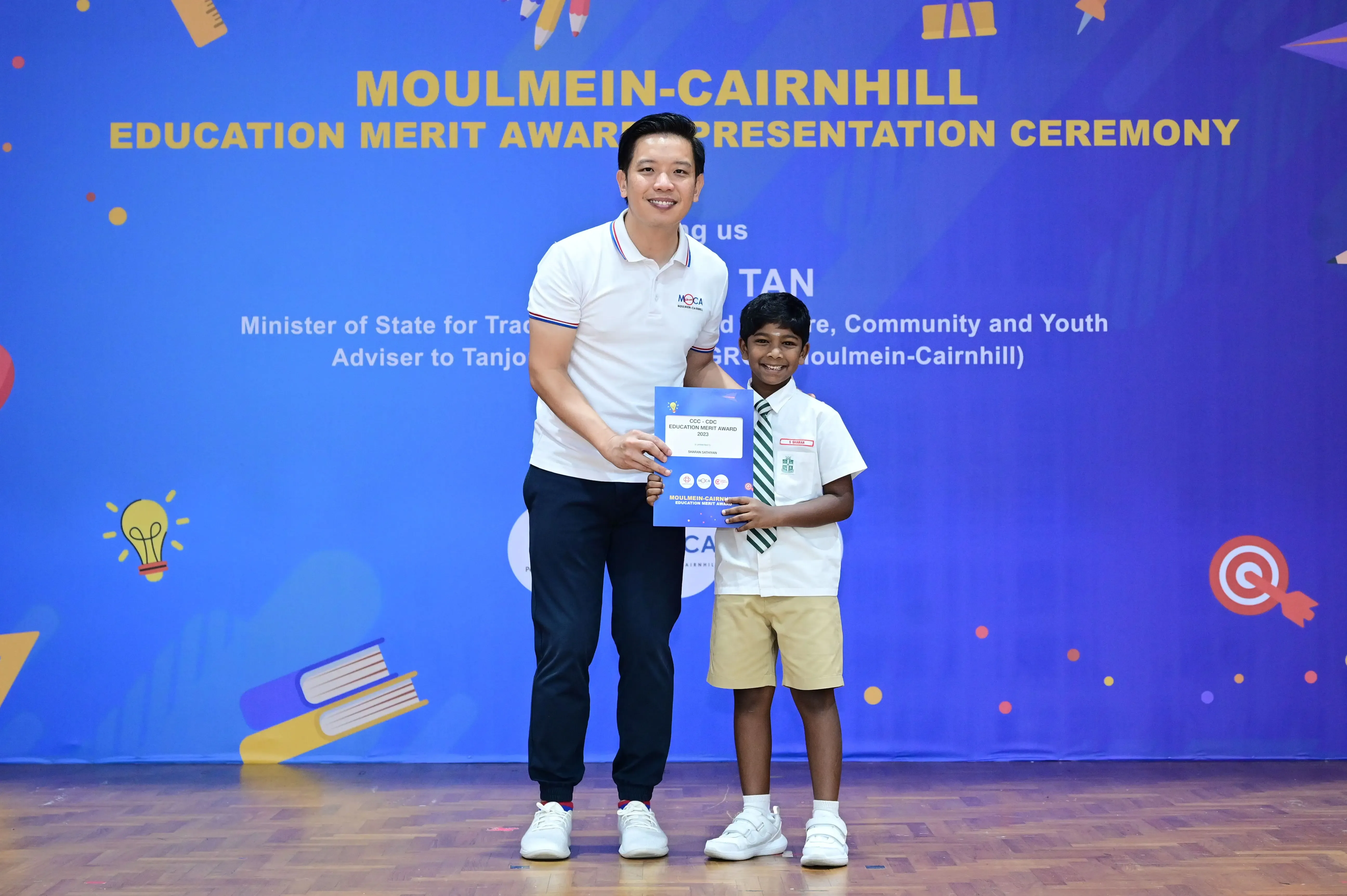 Man and young boy hold a blue award certificate on stage at Moulmein-Cairnhill Education Merit Award Ceremony.