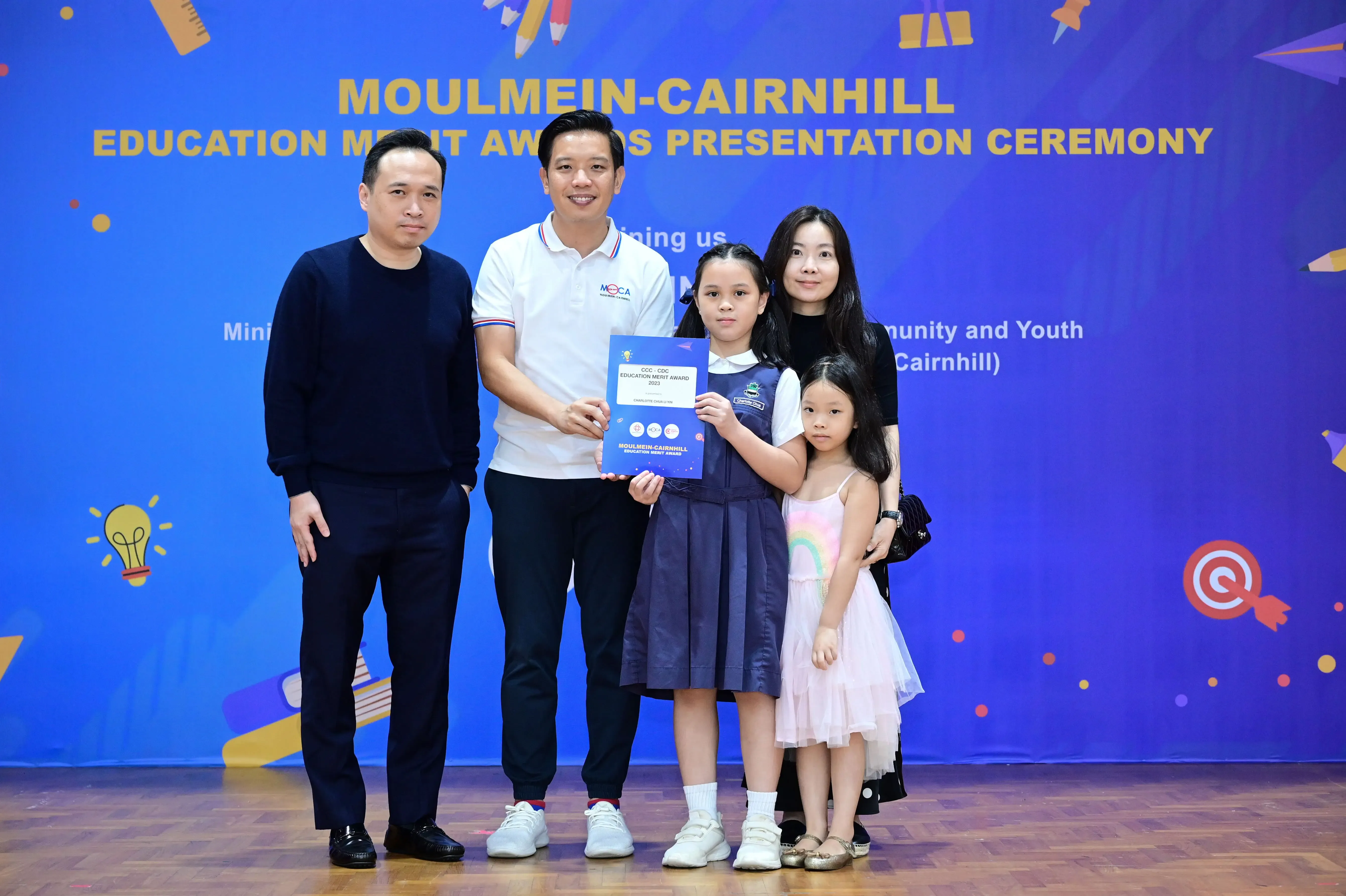 Five people pose before a Moulmein-Cairnhill Education Merit Awards banner, with a girl holding a certificate.