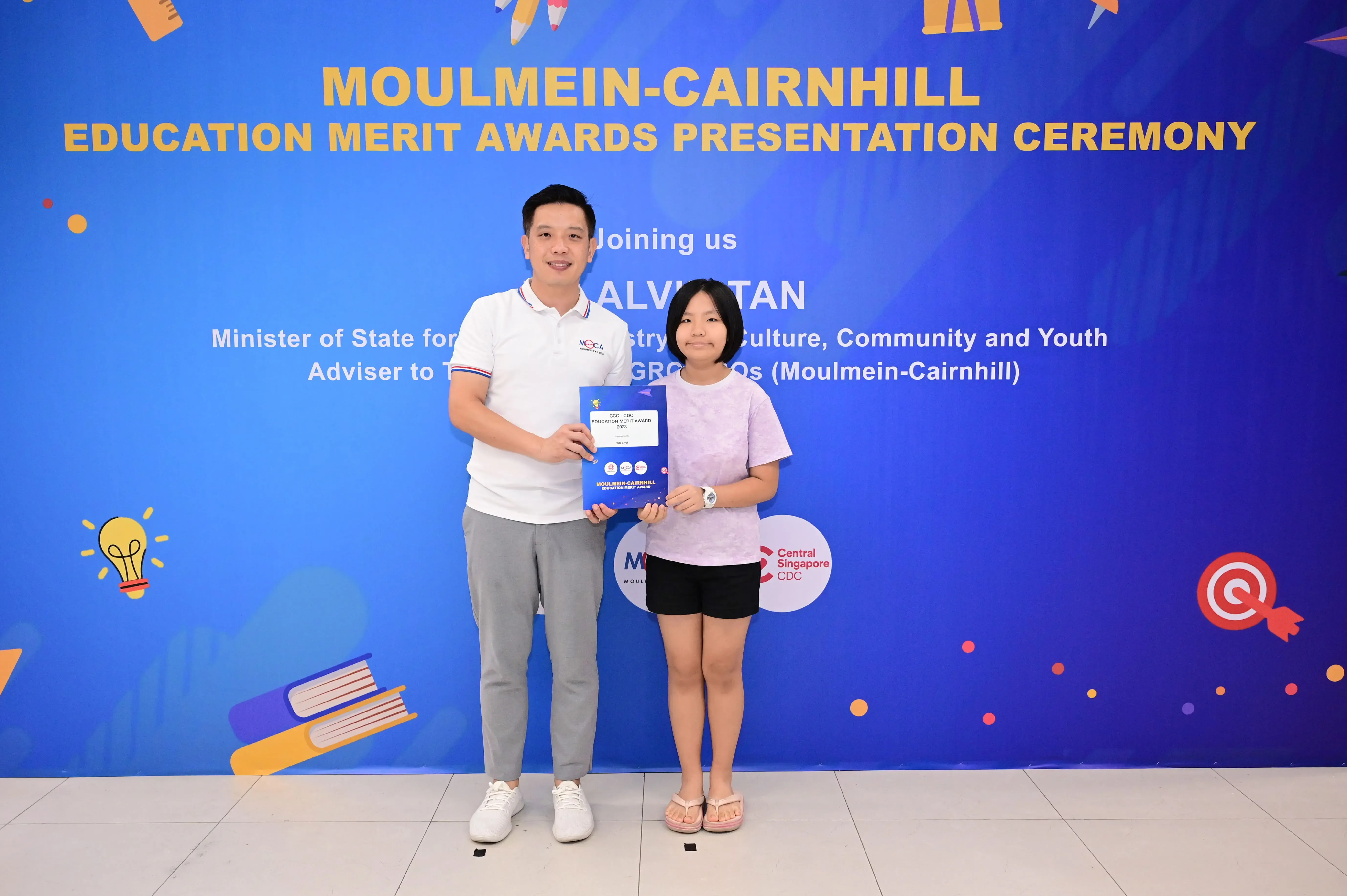 A man and a girl hold an award booklet in front of a Moulmein-Cairnhill Education Merit Awards banner.