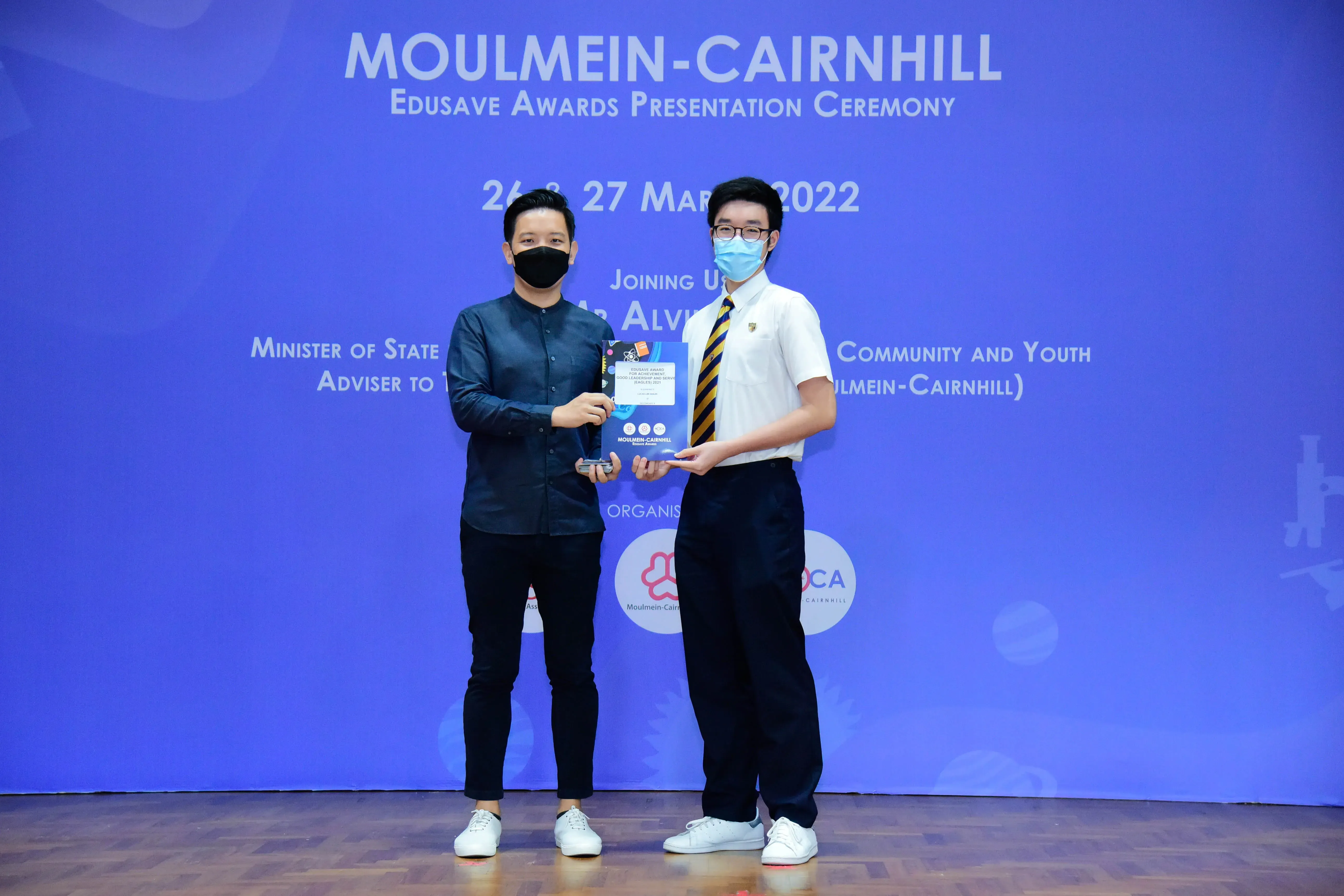 Two masked individuals hold an award at the Moulmein-Cairnhill Edusave Awards Presentation Ceremony, March 2022.