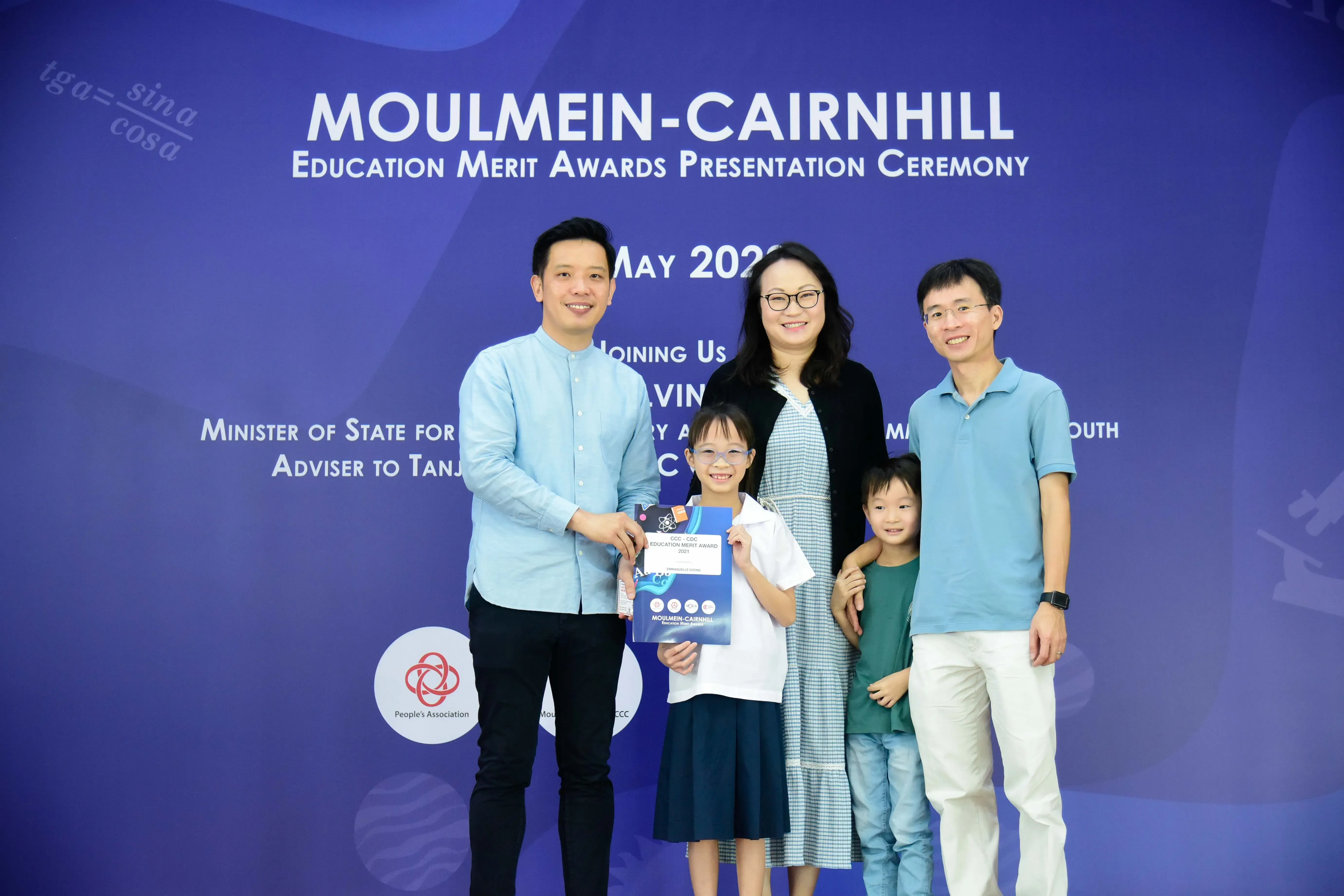 Four adults and two children pose at the Moulmein-Cairnhill Education Merit Awards Presentation Ceremony backdrop.