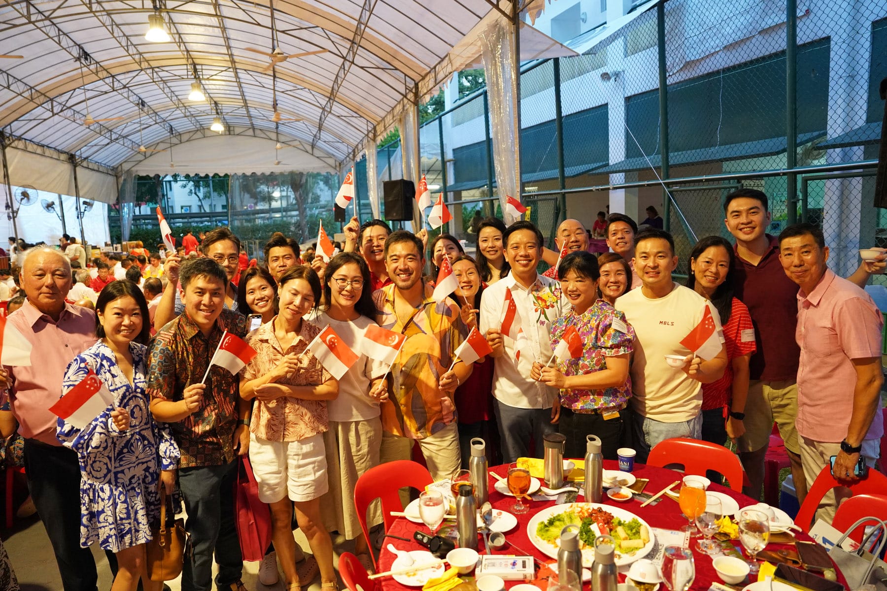 A large group of smiling people holding Singapore flags at an outdoor event under a white tent with food on tables.