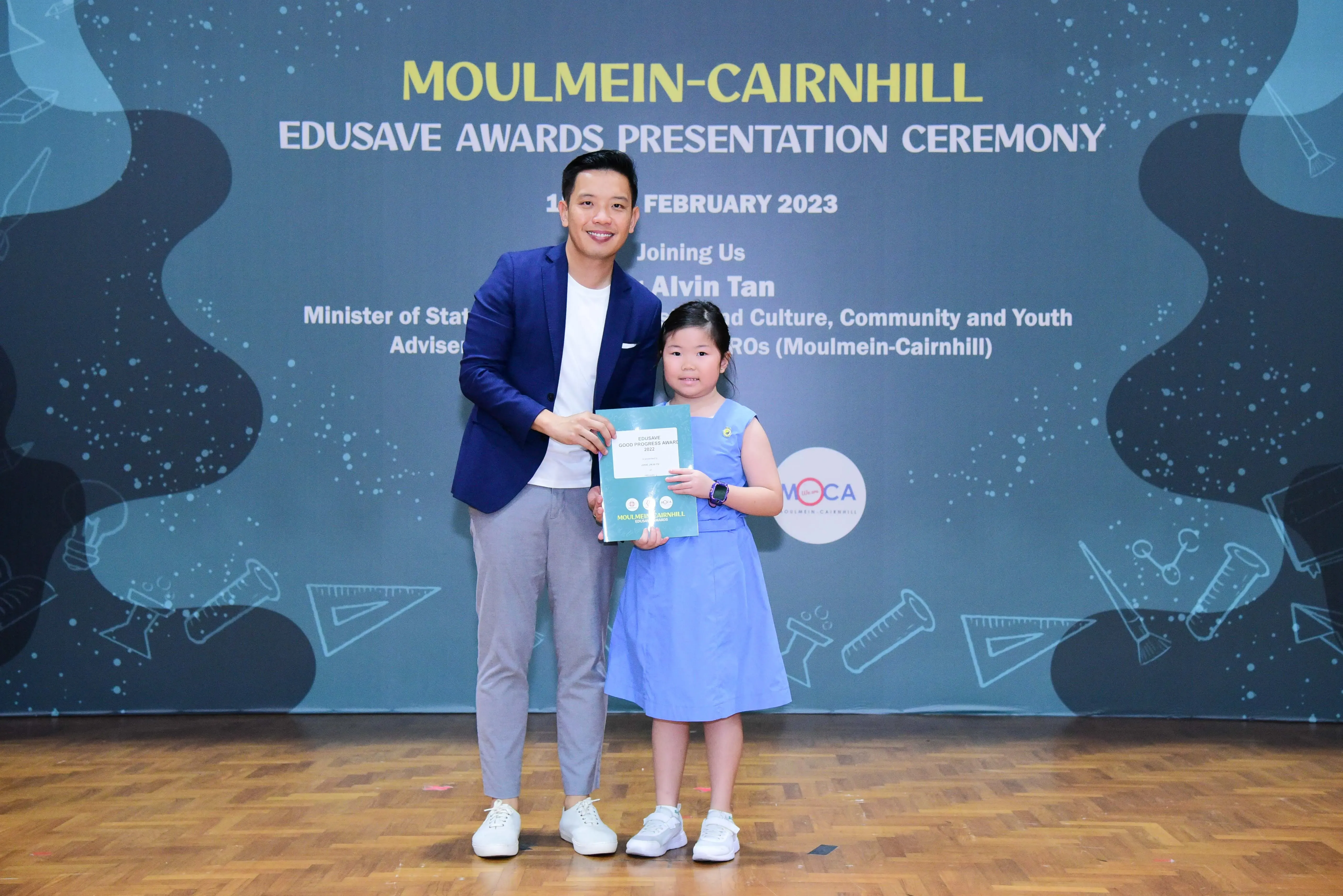 A man in a blue blazer and a young girl in a blue dress hold a certificate at the Moulmein-Cairnhill Edusave Awards 2023.