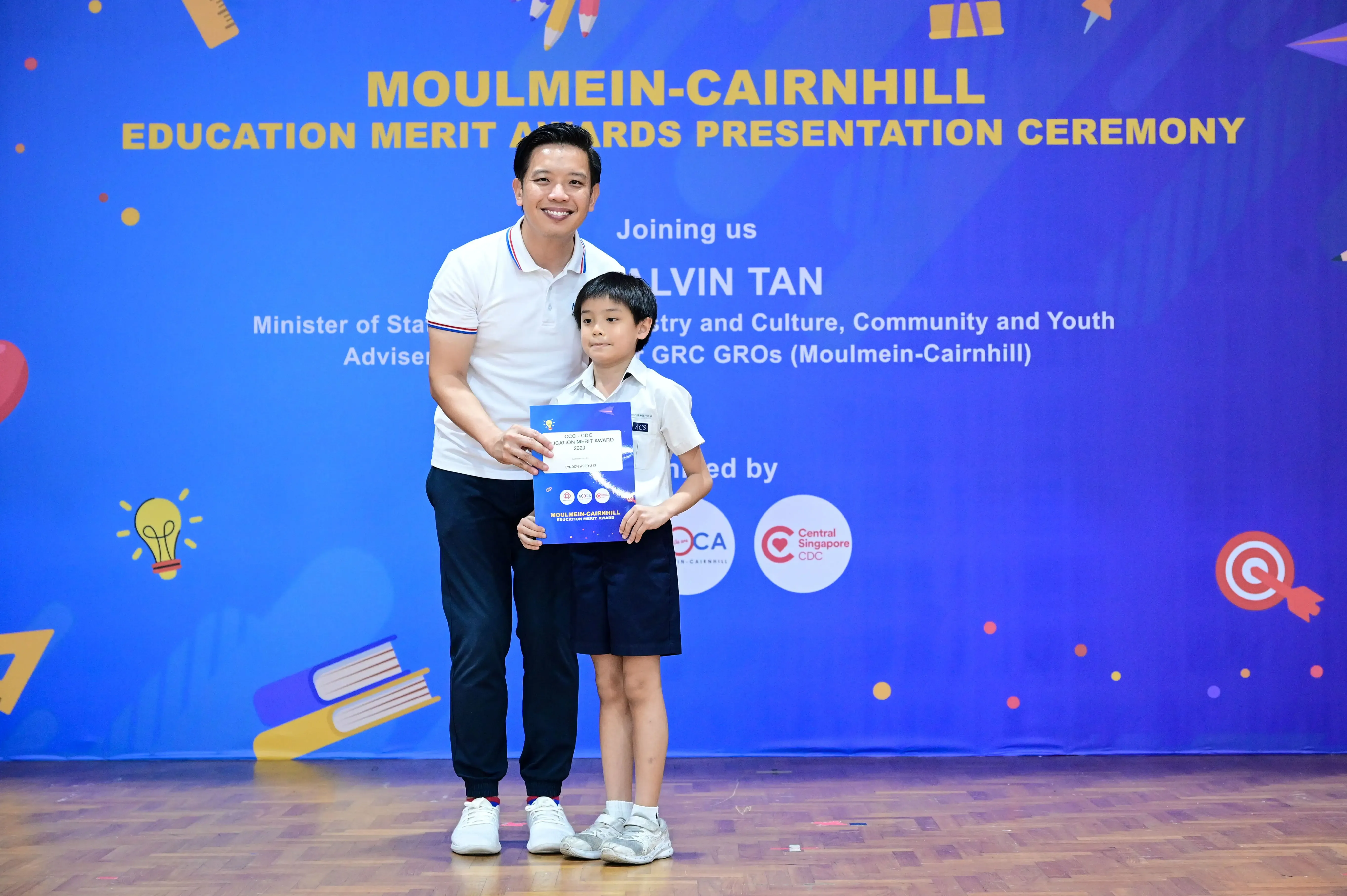A man and a young boy hold a certificate in front of a Moulmein-Cairnhill Education Merit Awards banner.