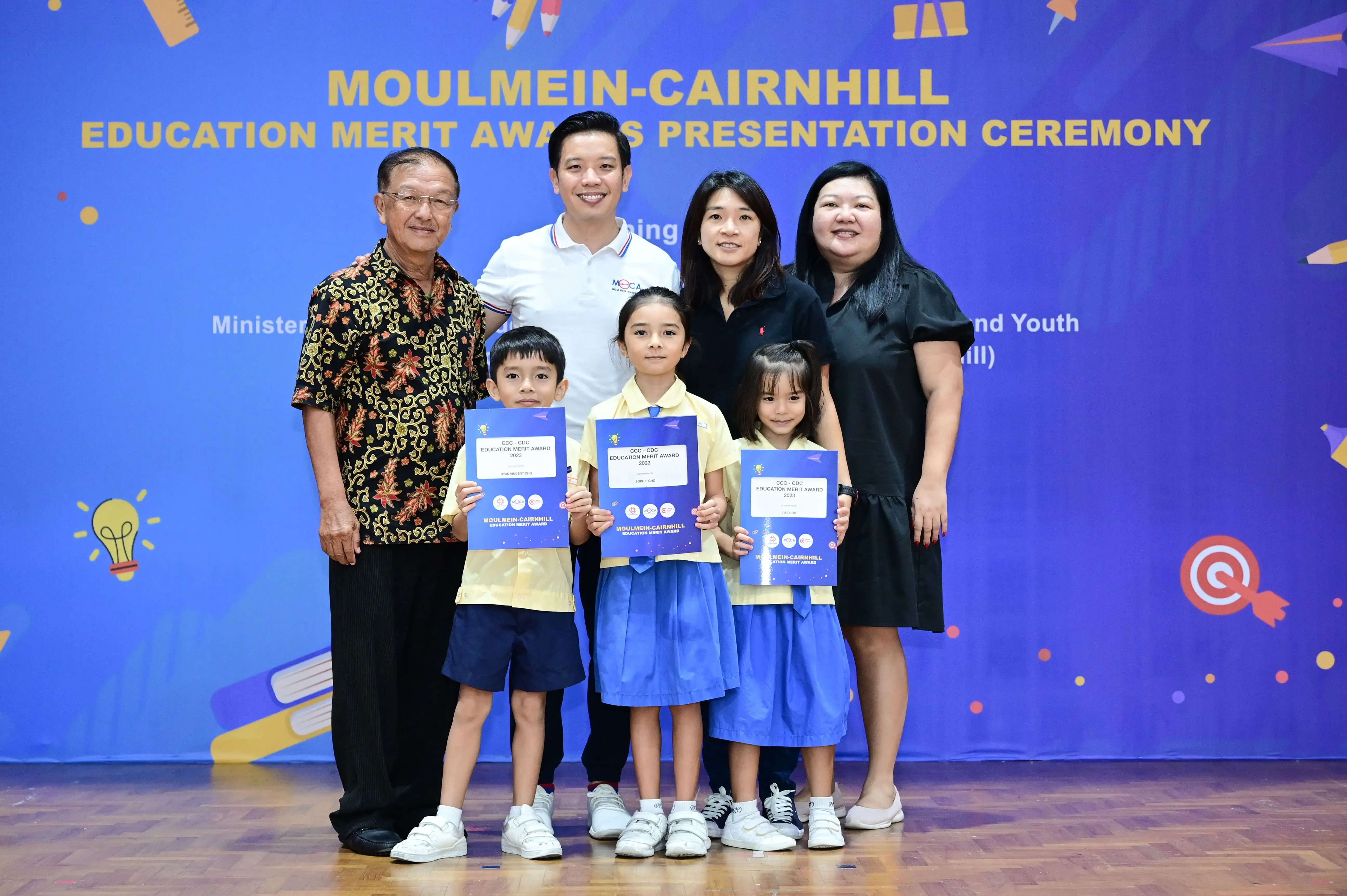 Three children hold blue award certificates at the Moulmein-Cairnhill Education Merit Awards Presentation Ceremony, flanked by four adults.